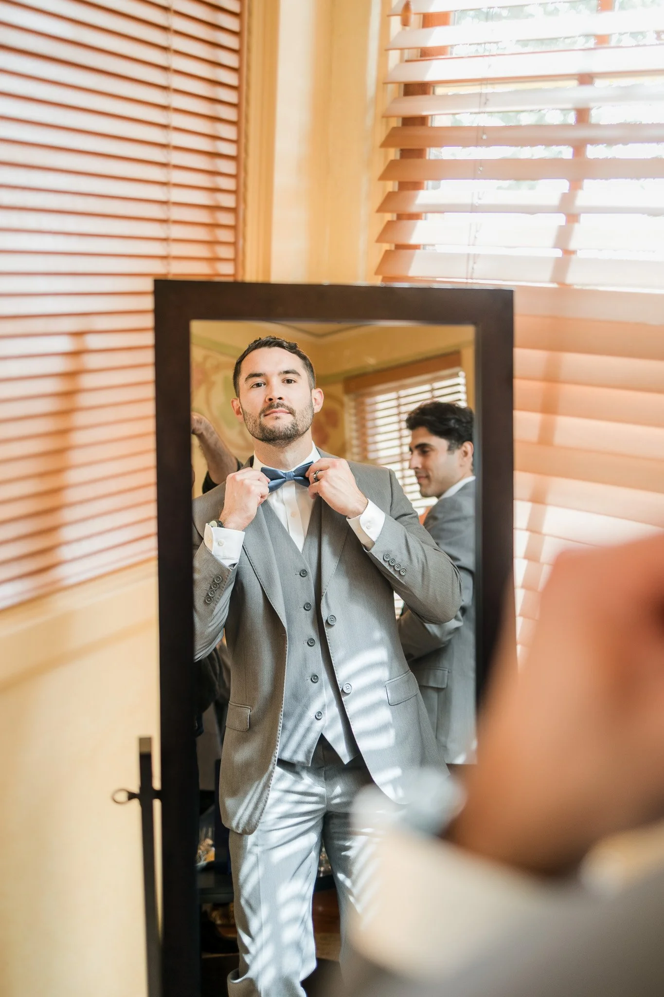 Man in gray suit adjusting a bow tie in front of a mirror, with another man in a matching suit in the background.