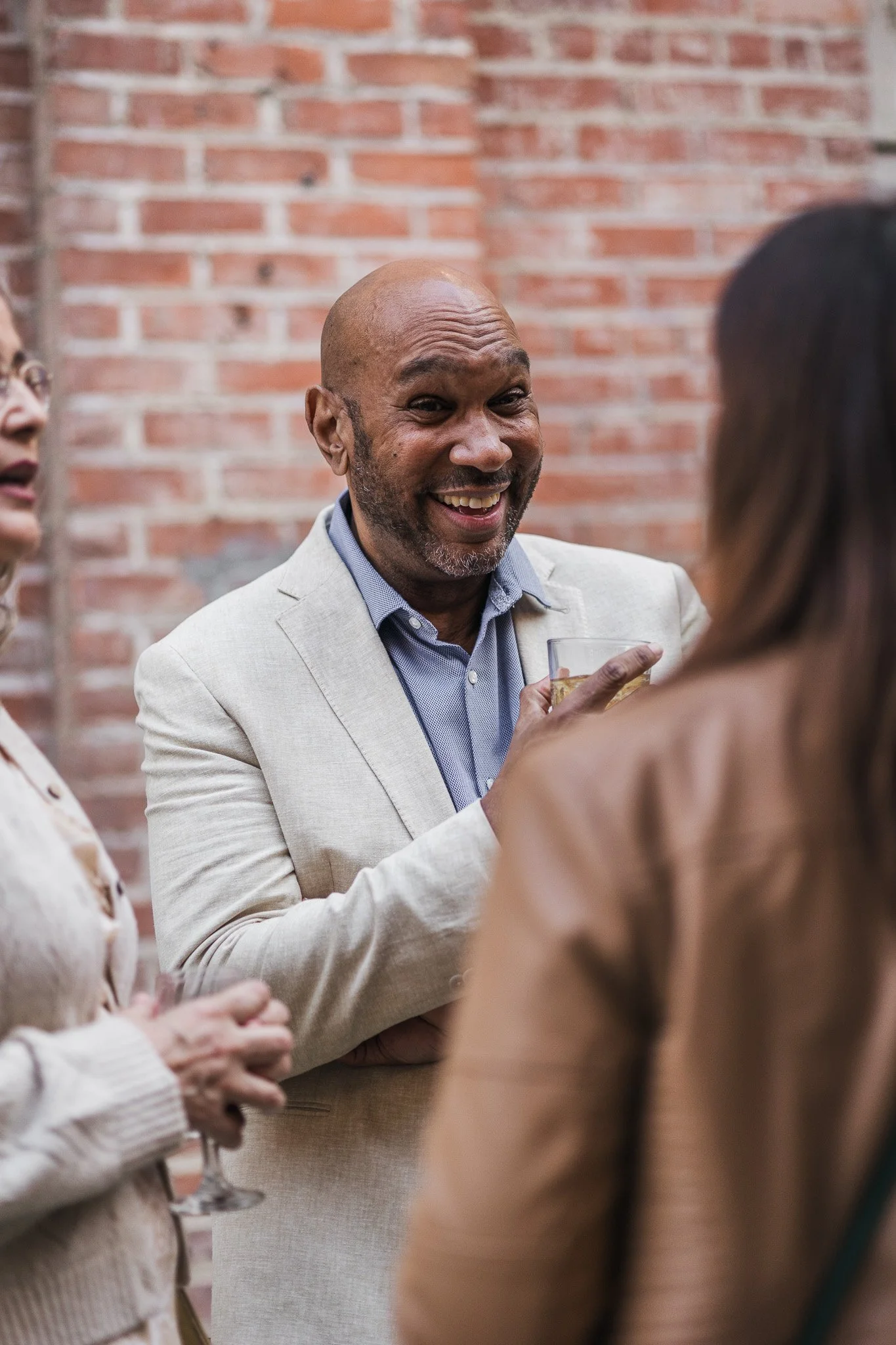 A man with a bald head and beard wearing a light-colored blazer and blue shirt, smiling and holding a glass of drink, talking to two women at a social gathering against a brick wall background.