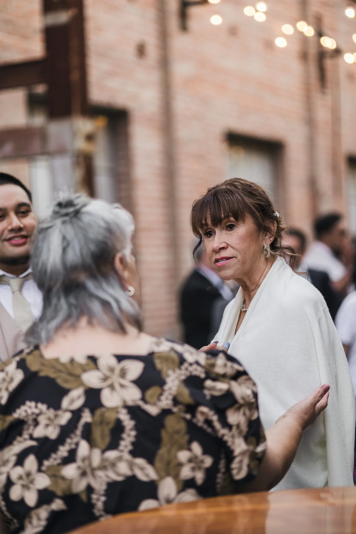 Three people engaged in conversation at an outdoor event, with a brick building and string lights in the background.
