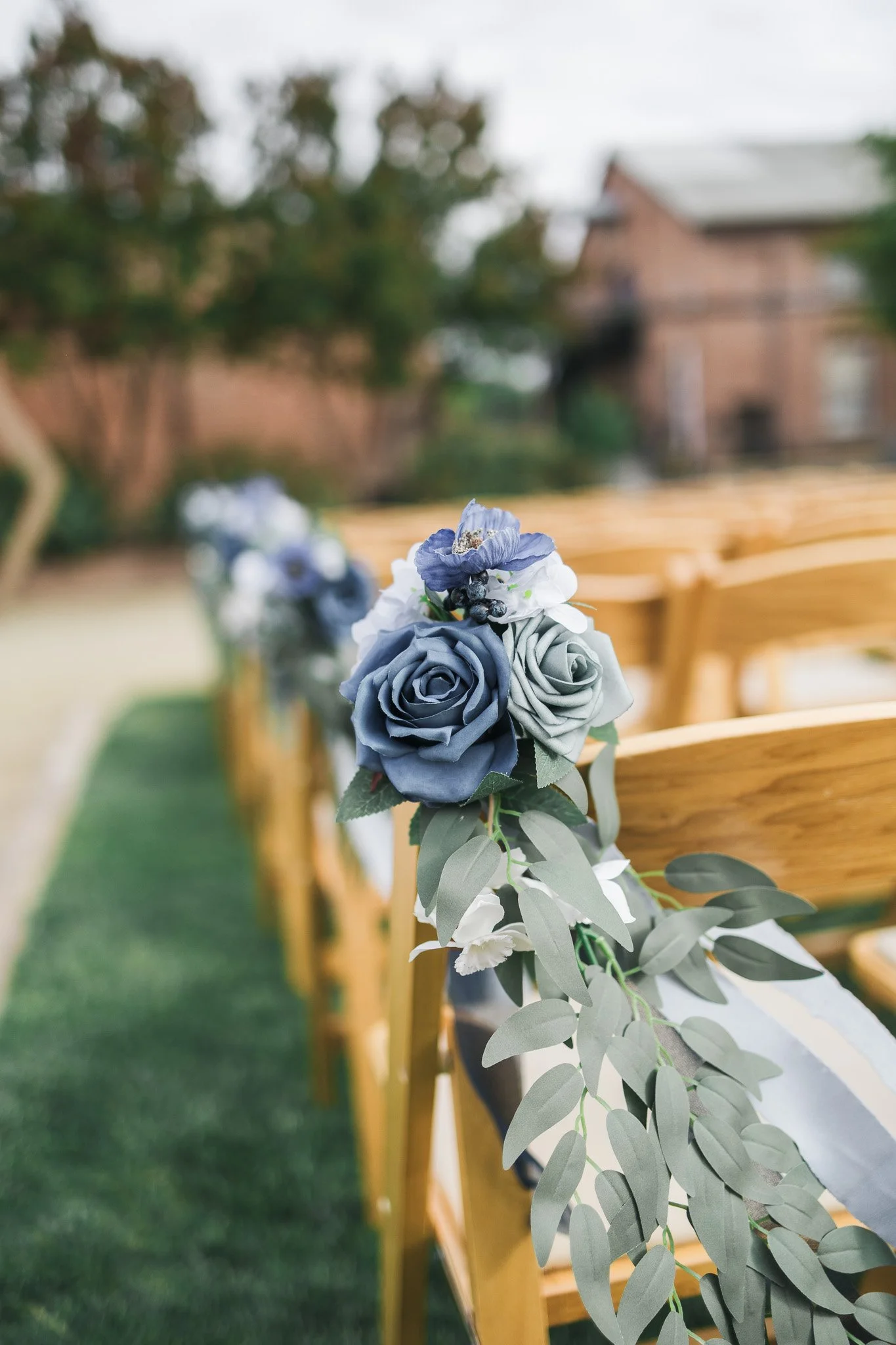 Close-up of a floral arrangement with blue and grey roses and greenery attached to the back of a wooden chair at an outdoor wedding ceremony.