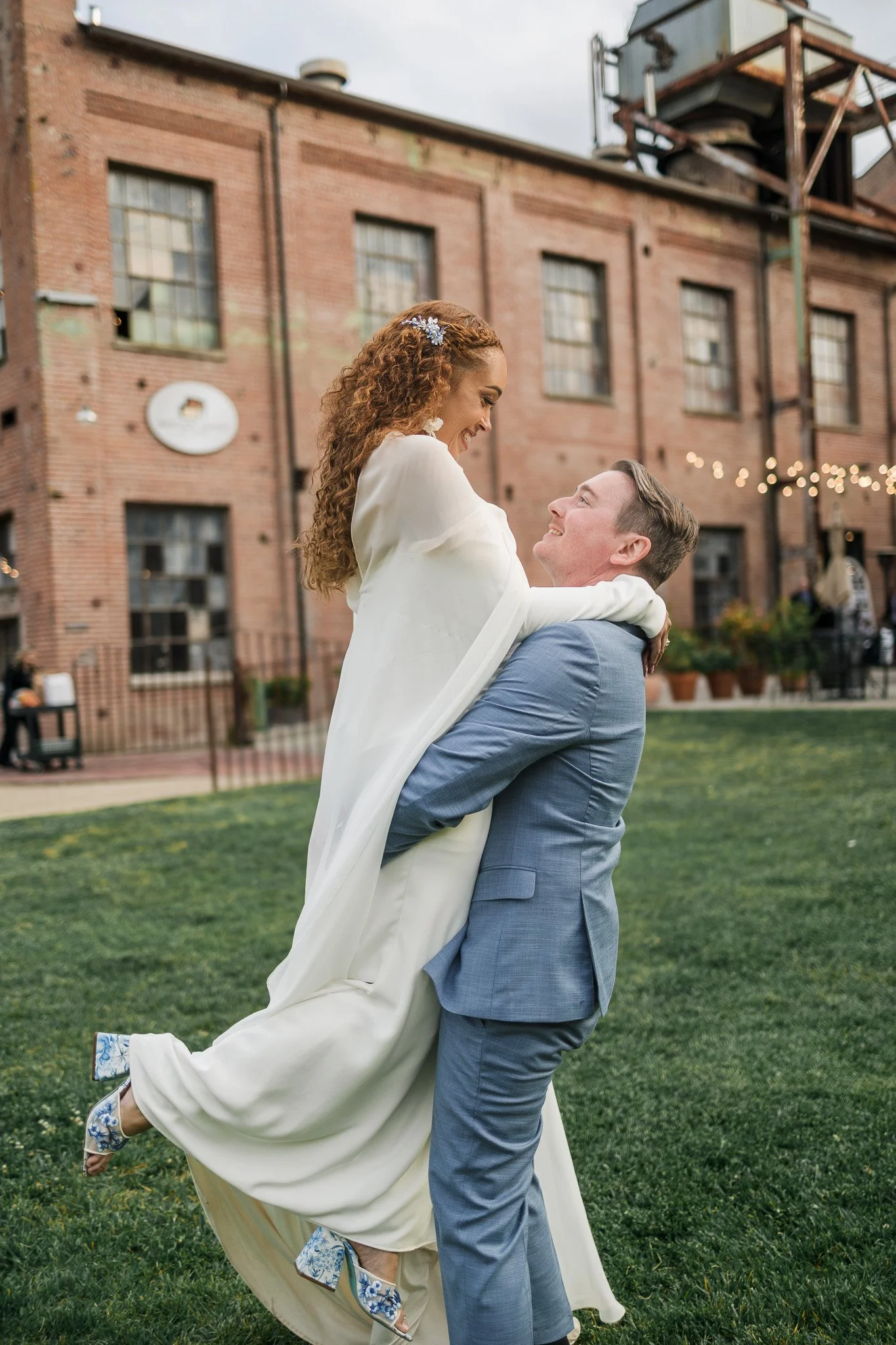 A man in a light grey suit lifts a woman in a white dress in a joyful embrace on a grassy area with an industrial brick building in the background.
