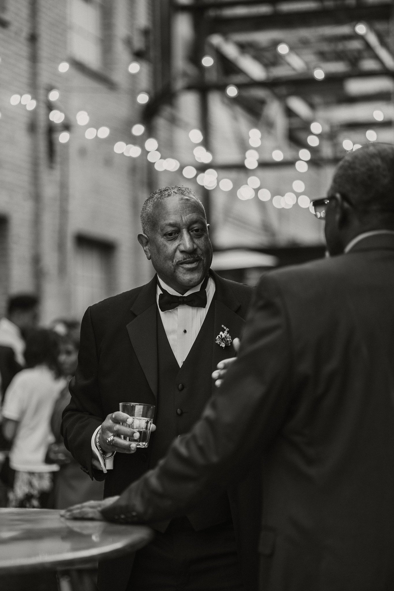 A black-and-white photo of two men in tuxedos conversing at a social event, with string lights and other guests in the background.