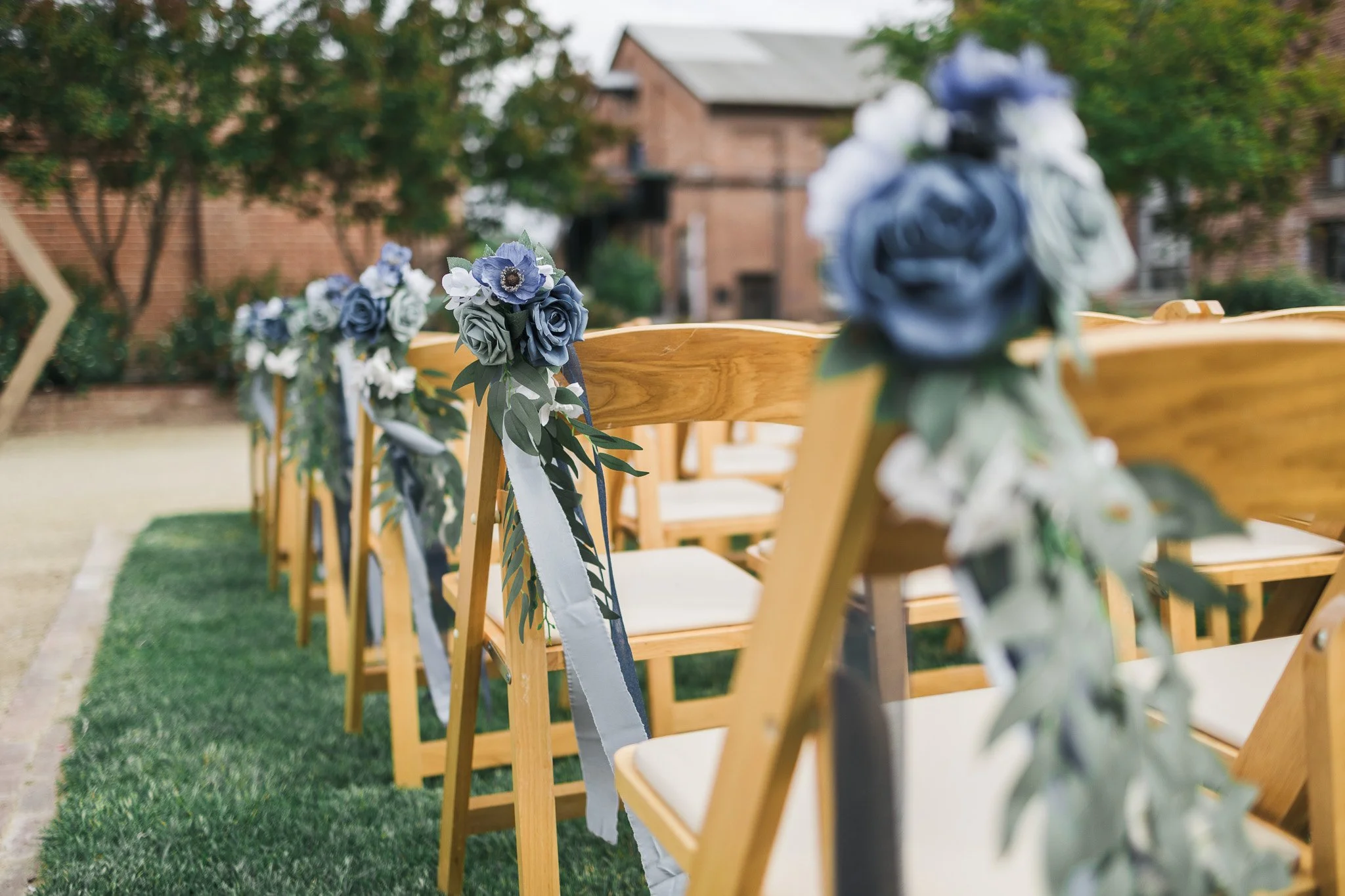 Wedding chairs decorated with blue and white floral arrangements and ribbons, set outside on a grassy area in front of a brick building.