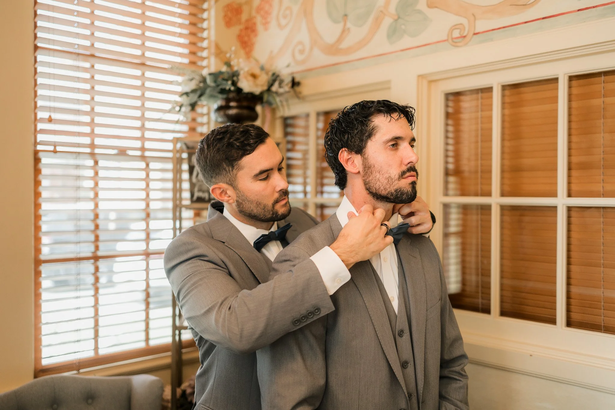 Two men in gray suits and black bow ties, one adjusting the other's collar in a room with wooden blinds and floral wall decor.