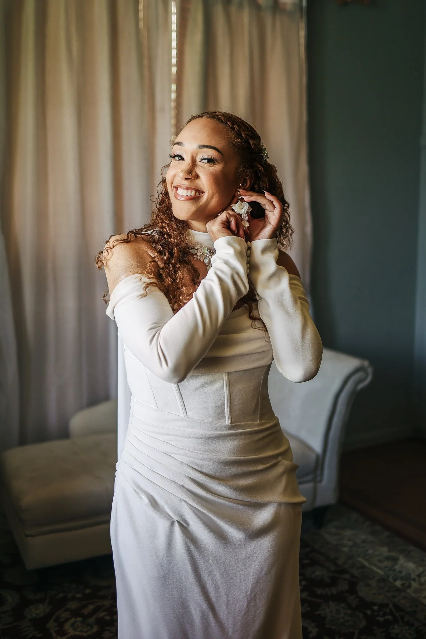 A woman in an elegant cream-colored dress is smiling while putting on earrings in a softly lit room with curtains and a cream-colored sofa.