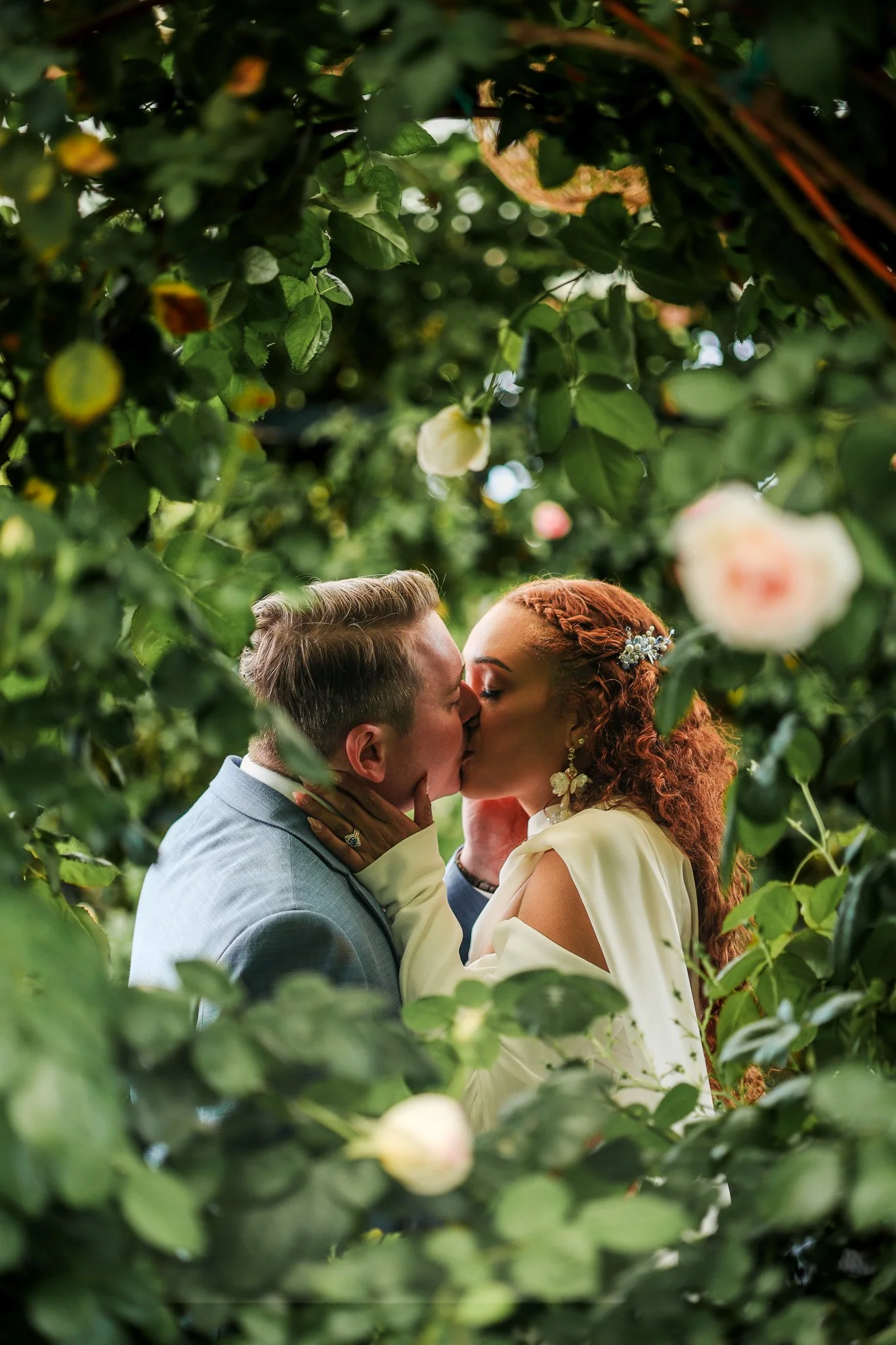 A couple kissing amidst lush greenery and blooming flowers.