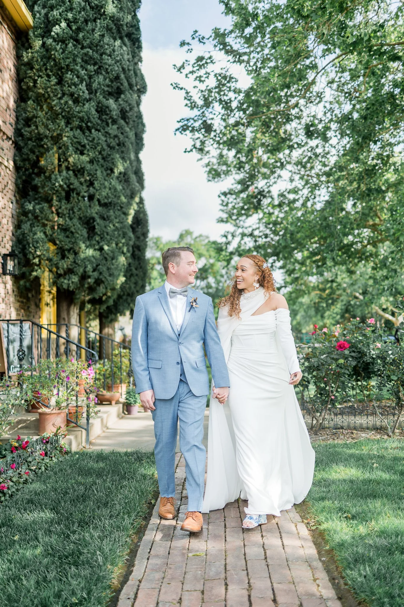 A bride and groom walking hand in hand on a brick path outdoors, smiling at each other, with lush greenery and blooming flowers around them.