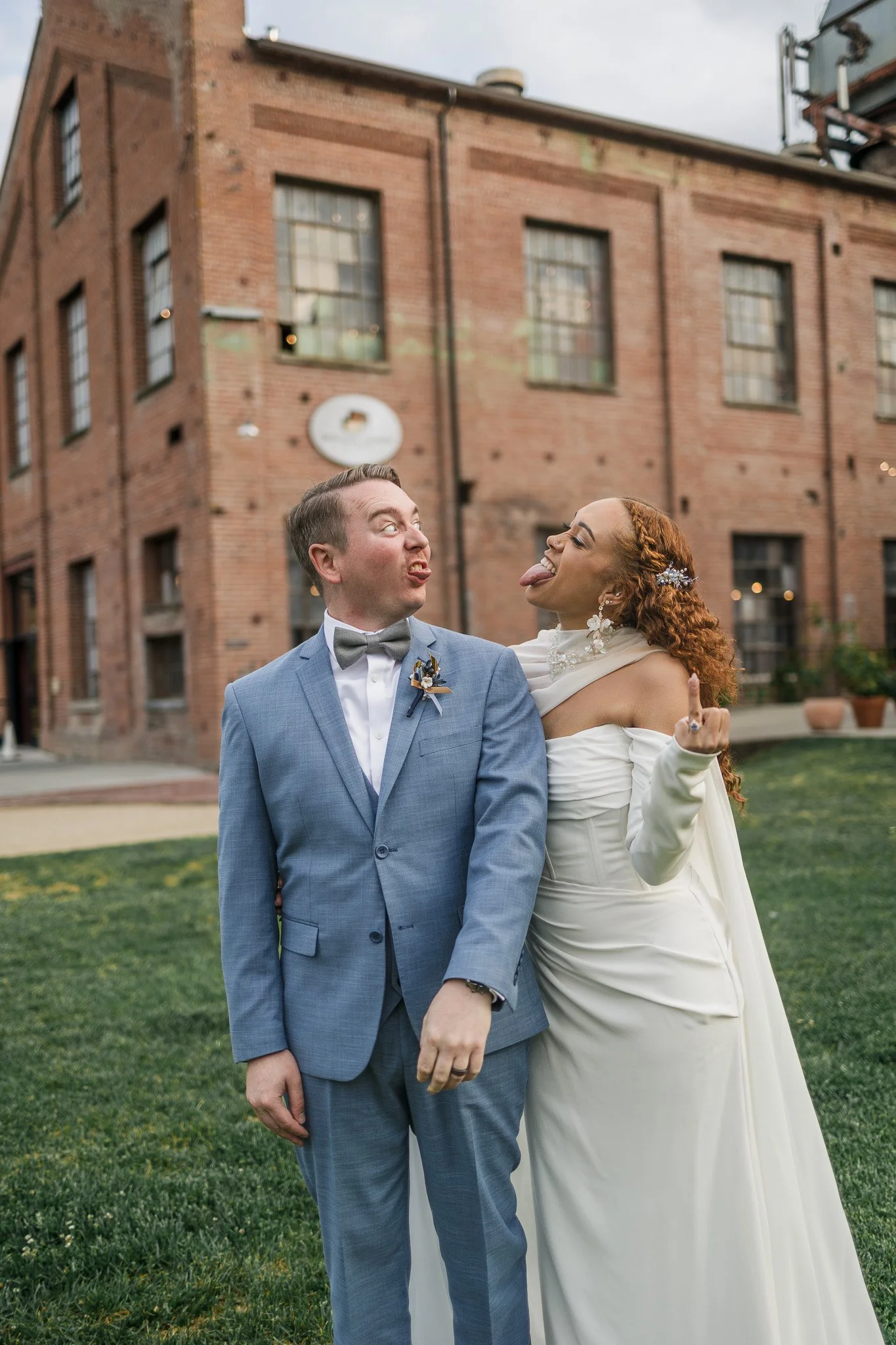 A couple in wedding attire making goofy faces and sticking out their tongues, standing outdoors in front of a brick building.