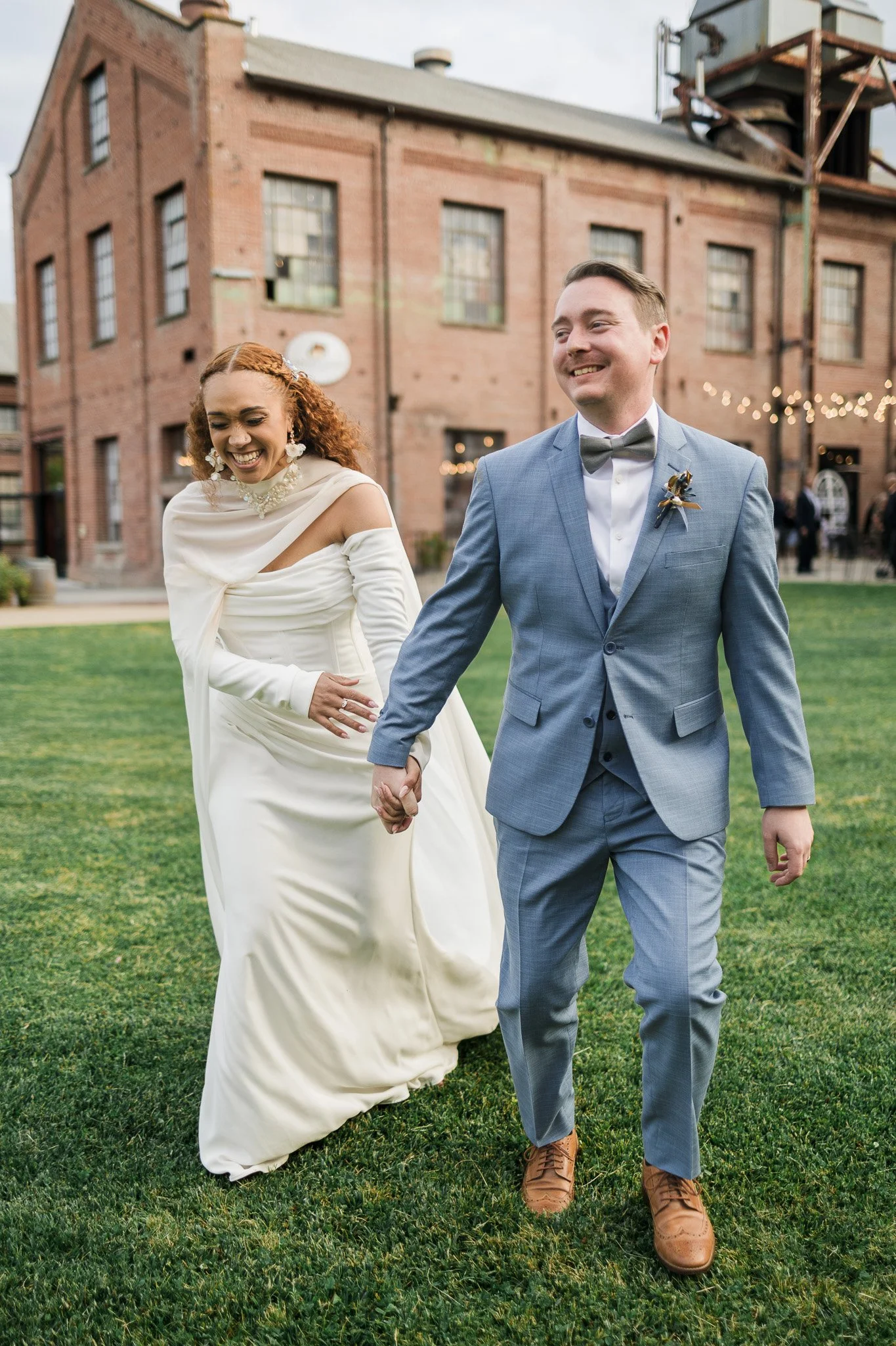 A bride and groom walking hand in hand on a grassy outdoor area, smiling and laughing. The bride is in a white wedding dress with a draped neckline and the groom is in a blue suit with a bow tie. Behind them is a brick building with string lights.