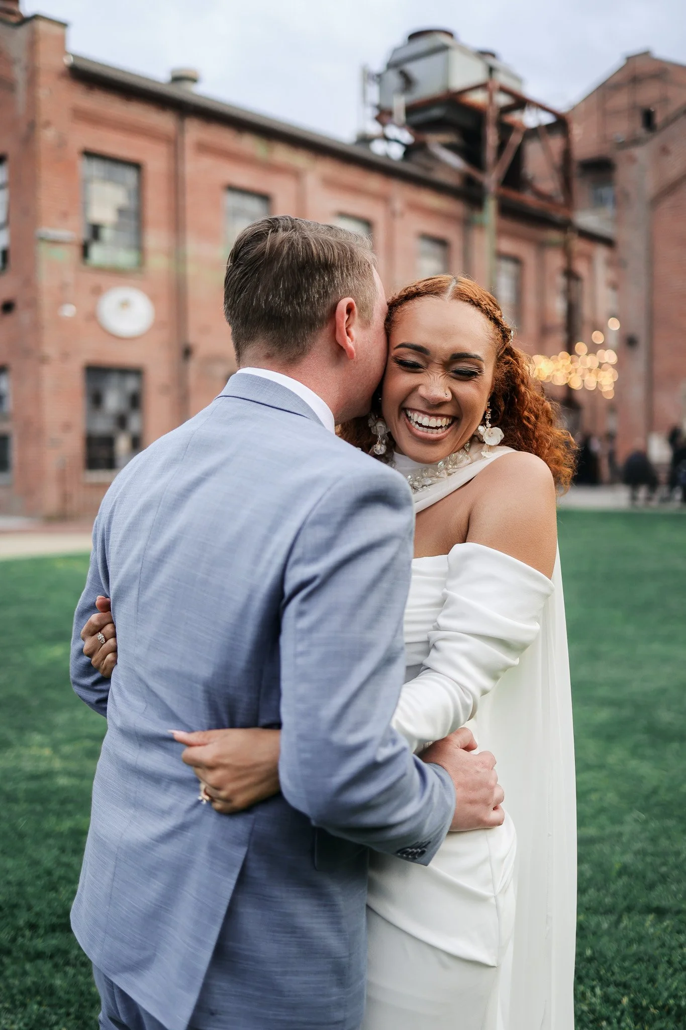 A joyous bride and groom sharing a hug outdoors on a grassy area with red brick buildings in the background, during their wedding celebration.