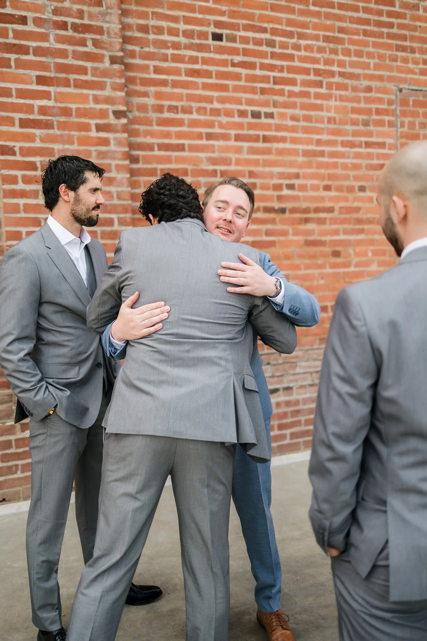 Four men in suits are standing against a brick wall, with two of them hugging and the others observing. One man with a beard and dark hair is standing with his hands in his pockets, watching the scene. All are dressed in formal grey suits.