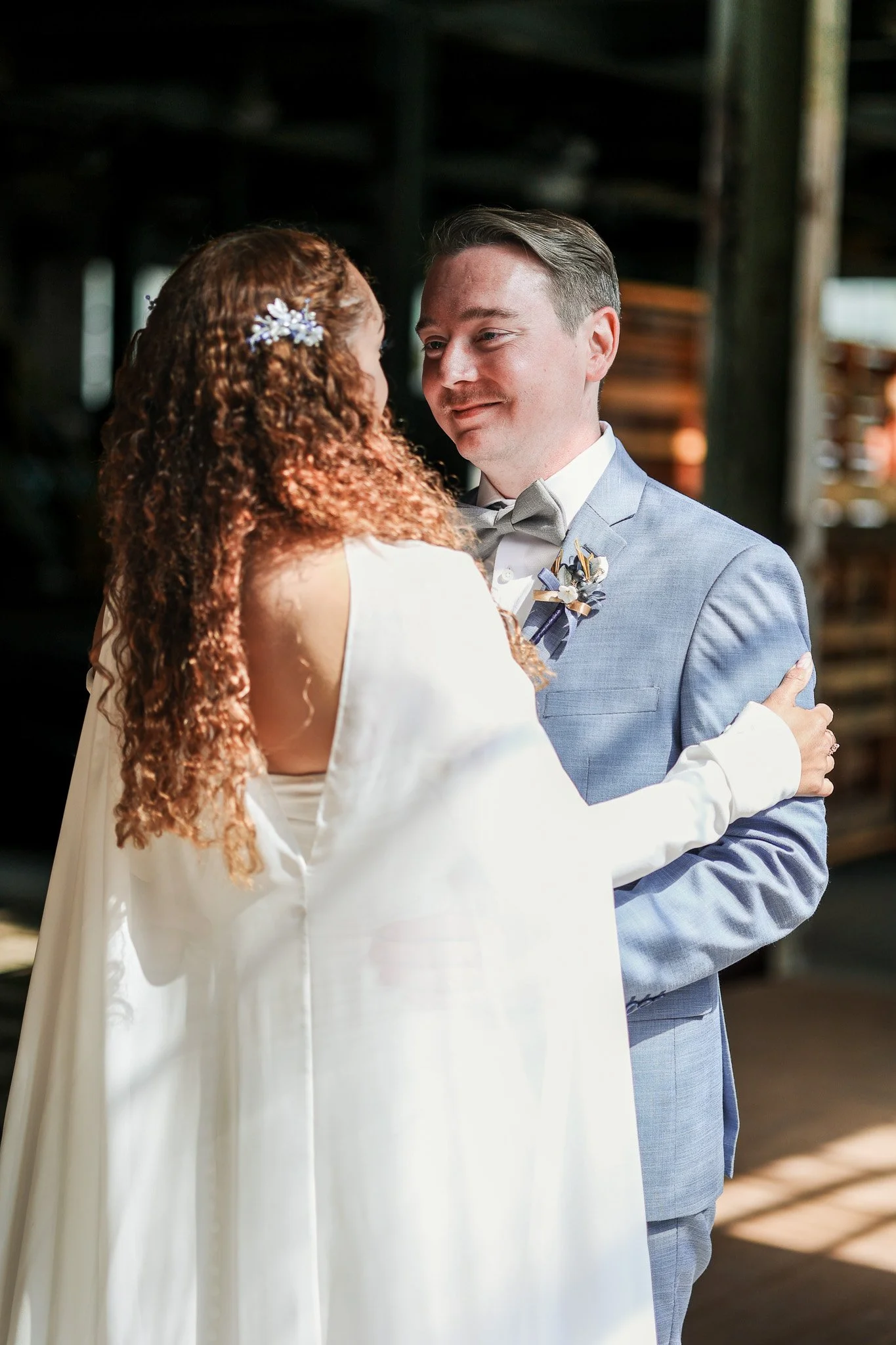 A couple, dressed in wedding attire, sharing a moment indoors with natural light, wooden background, and warm expressions.