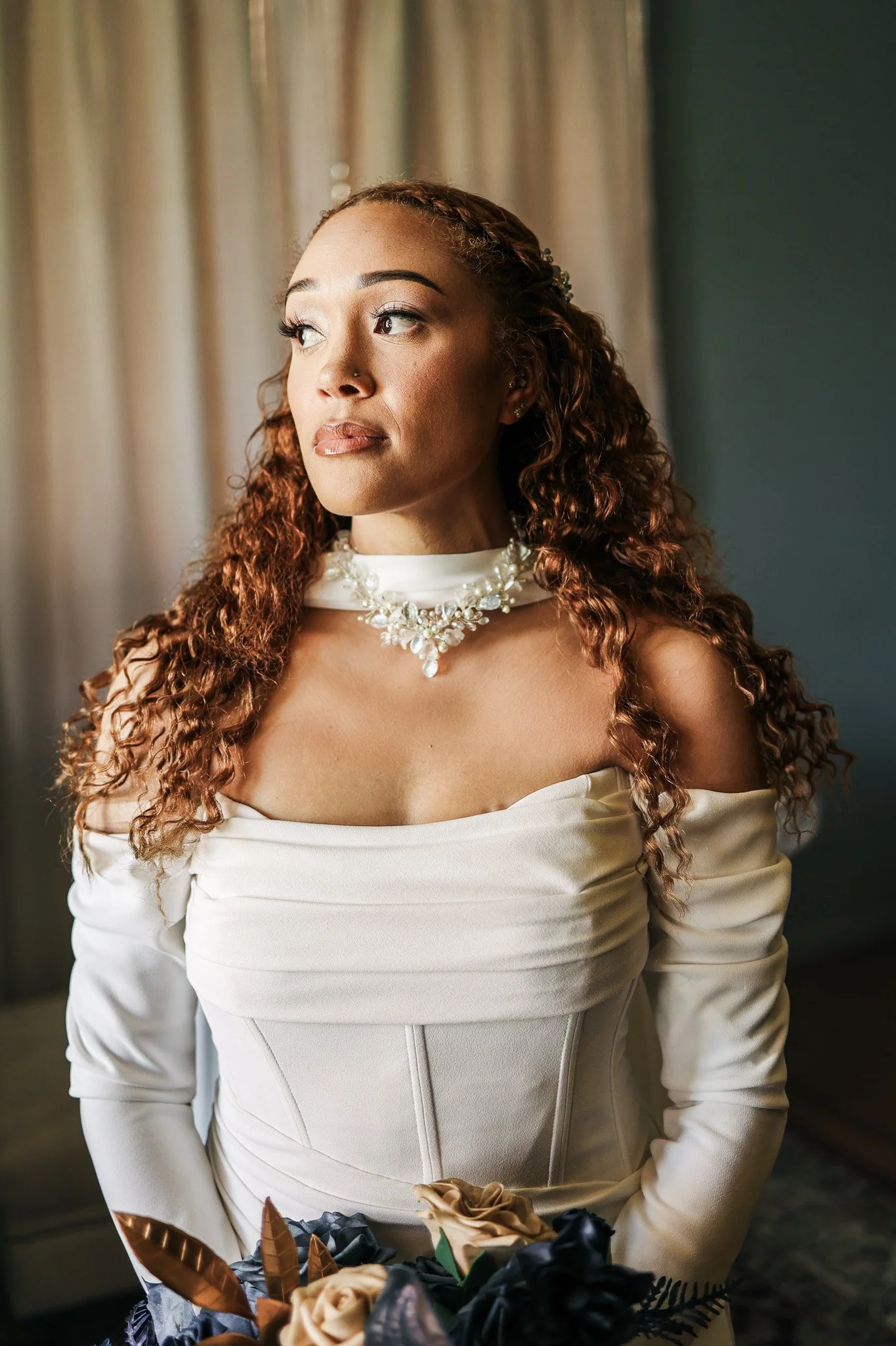 A woman with long, curly reddish hair and light makeup, wearing a pale off-shoulder dress with long sleeves, a white choker with pearl and gem embellishments, and holding a bouquet of flowers, standing near a window with light-colored curtains.