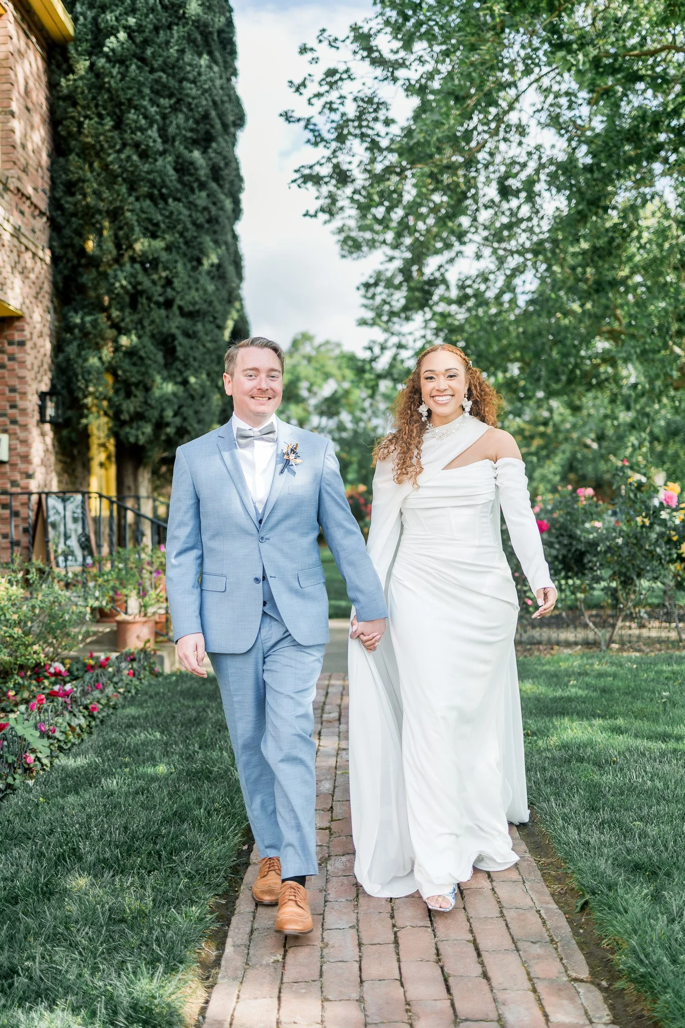A newlywed couple walking hand in hand on a brick pathway in a garden, smiling, with greenery and flowers around.