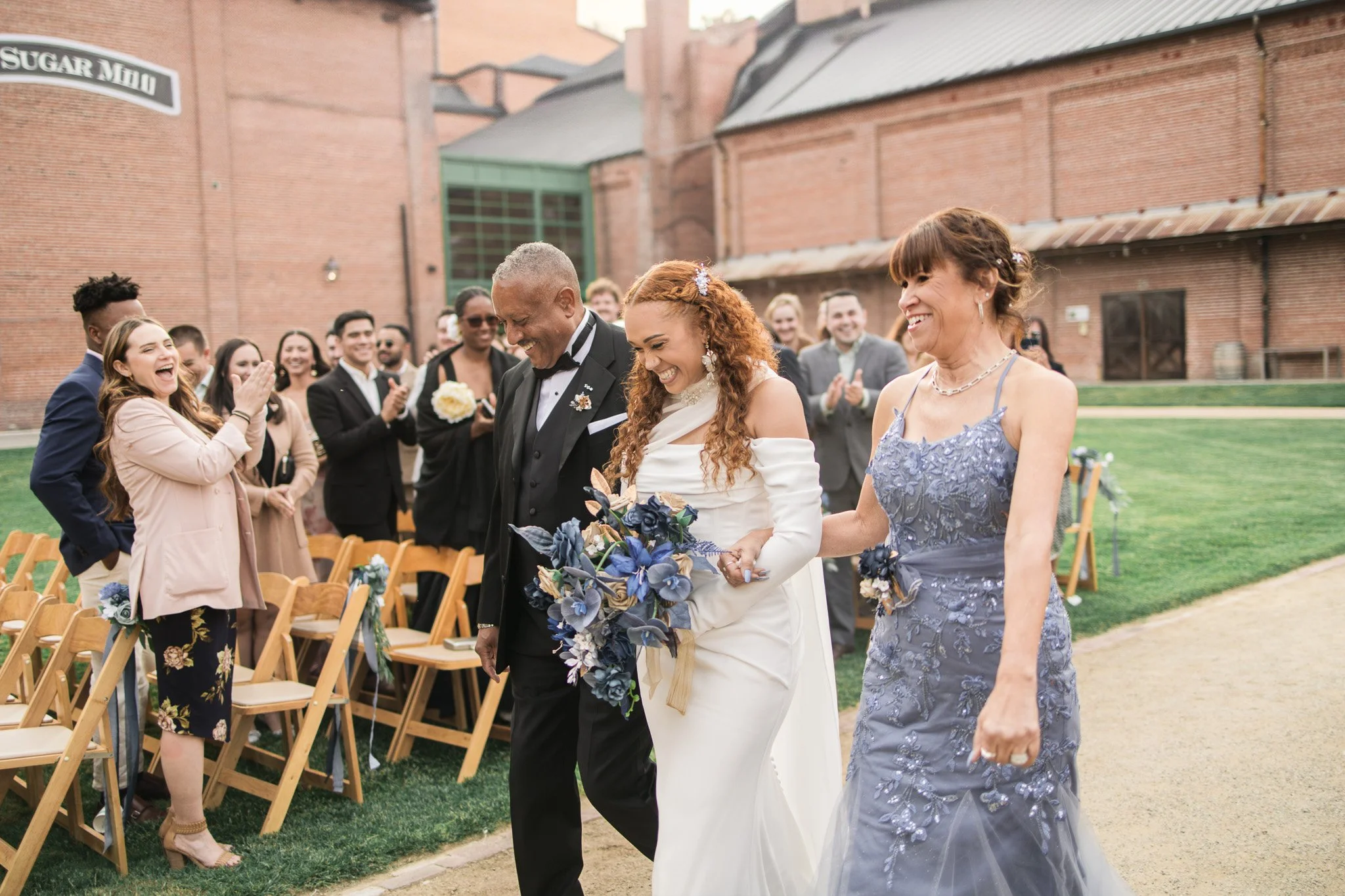 A bride in a white wedding dress holding a bouquet, walking next to a man in a black tuxedo and a woman in a gray lace dress, with guests applauding and smiling in the background at an outdoor wedding ceremony.