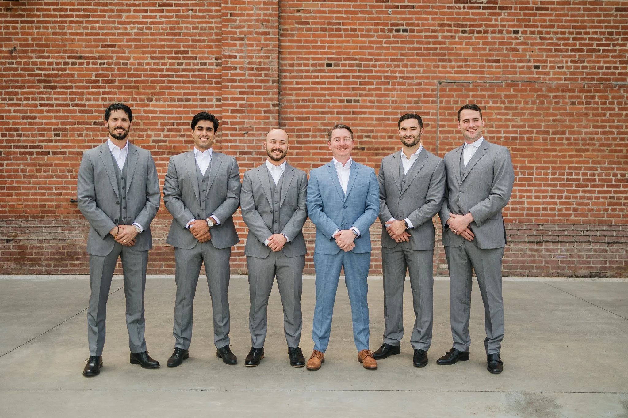 Six men dressed in suits standing in a line against a brick wall, smiling for a photo.
