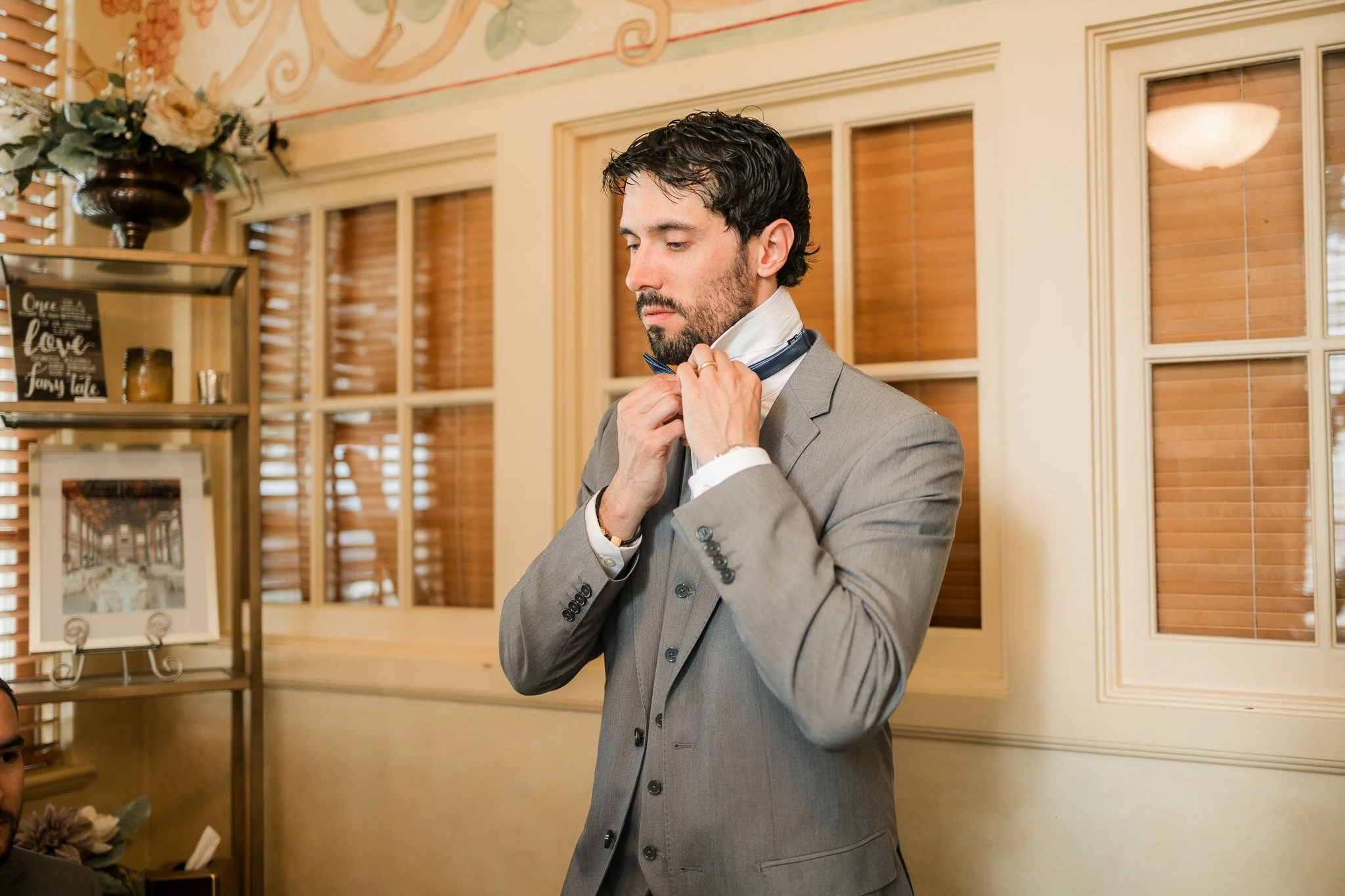 A man in a gray suit adjusting his bow tie in a warmly decorated room with wooden shutters and framed pictures.