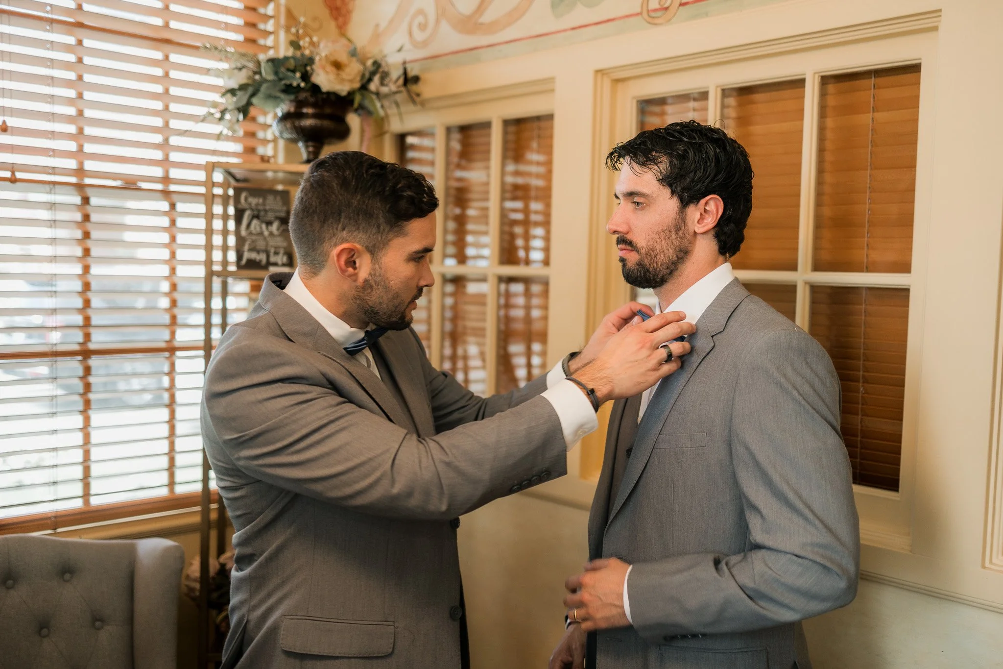 Two men in gray suits preparing for a formal event indoors, with one adjusting the other's bowtie.