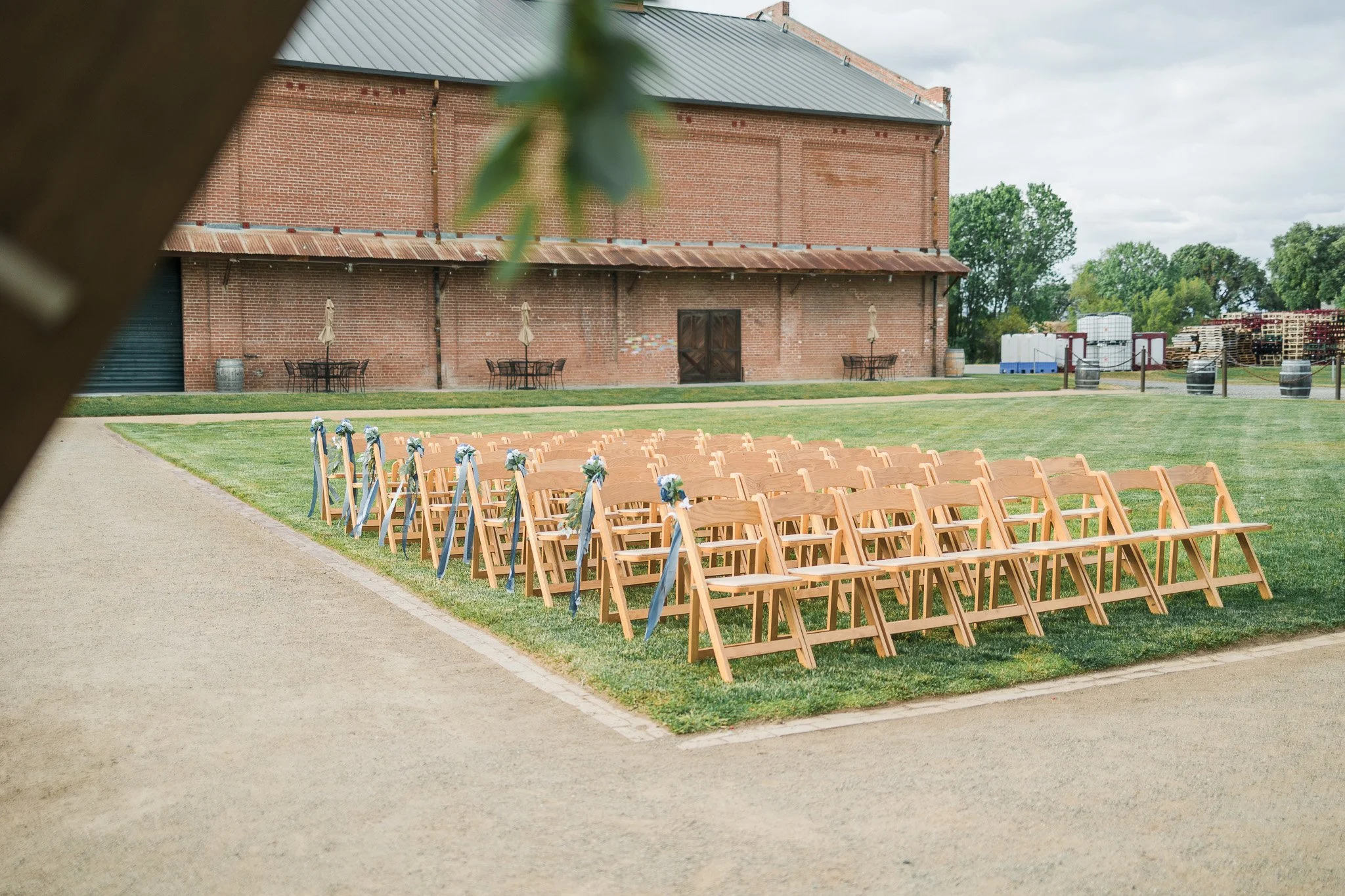 Outdoor wedding ceremony setup with rows of wooden chairs decorated with blue ribbons and white flowers, on a grassy area near a rustic brick building.