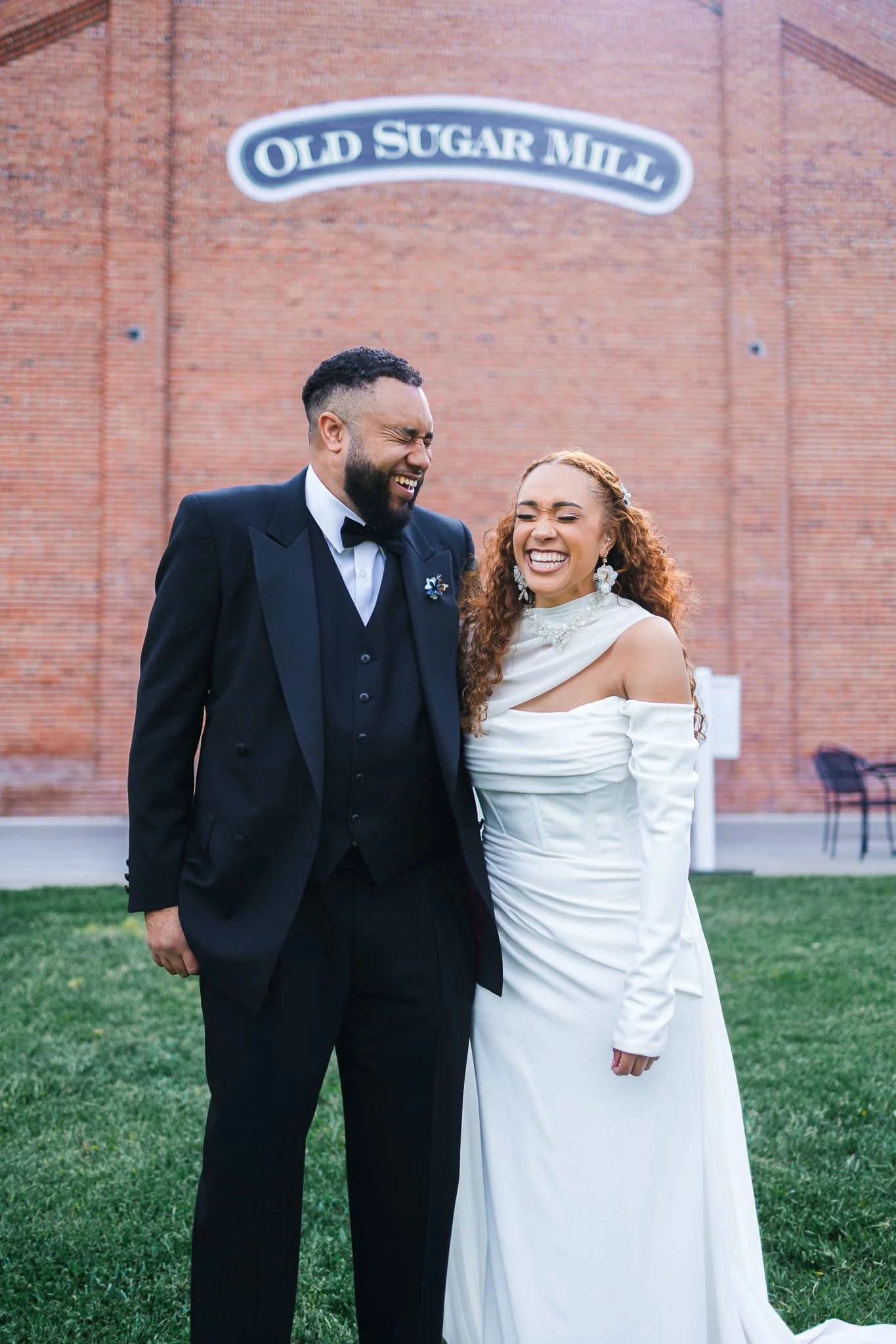 A joyful couple in wedding attire laughing in front of a brick wall with a sign that reads "Old Sugar Mill."