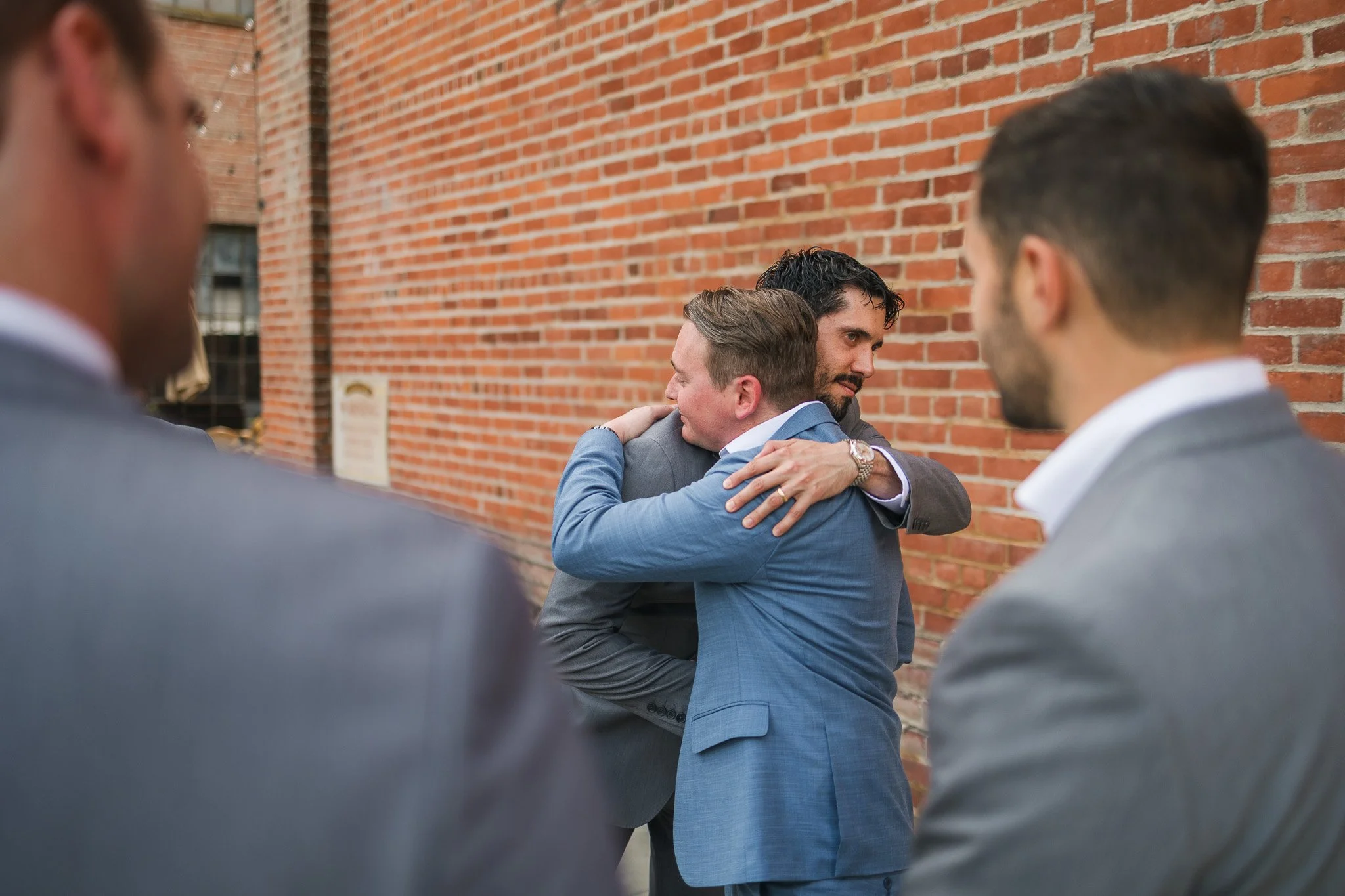 Two men in suits hugging each other outside against a brick wall while two other men in suits look on.