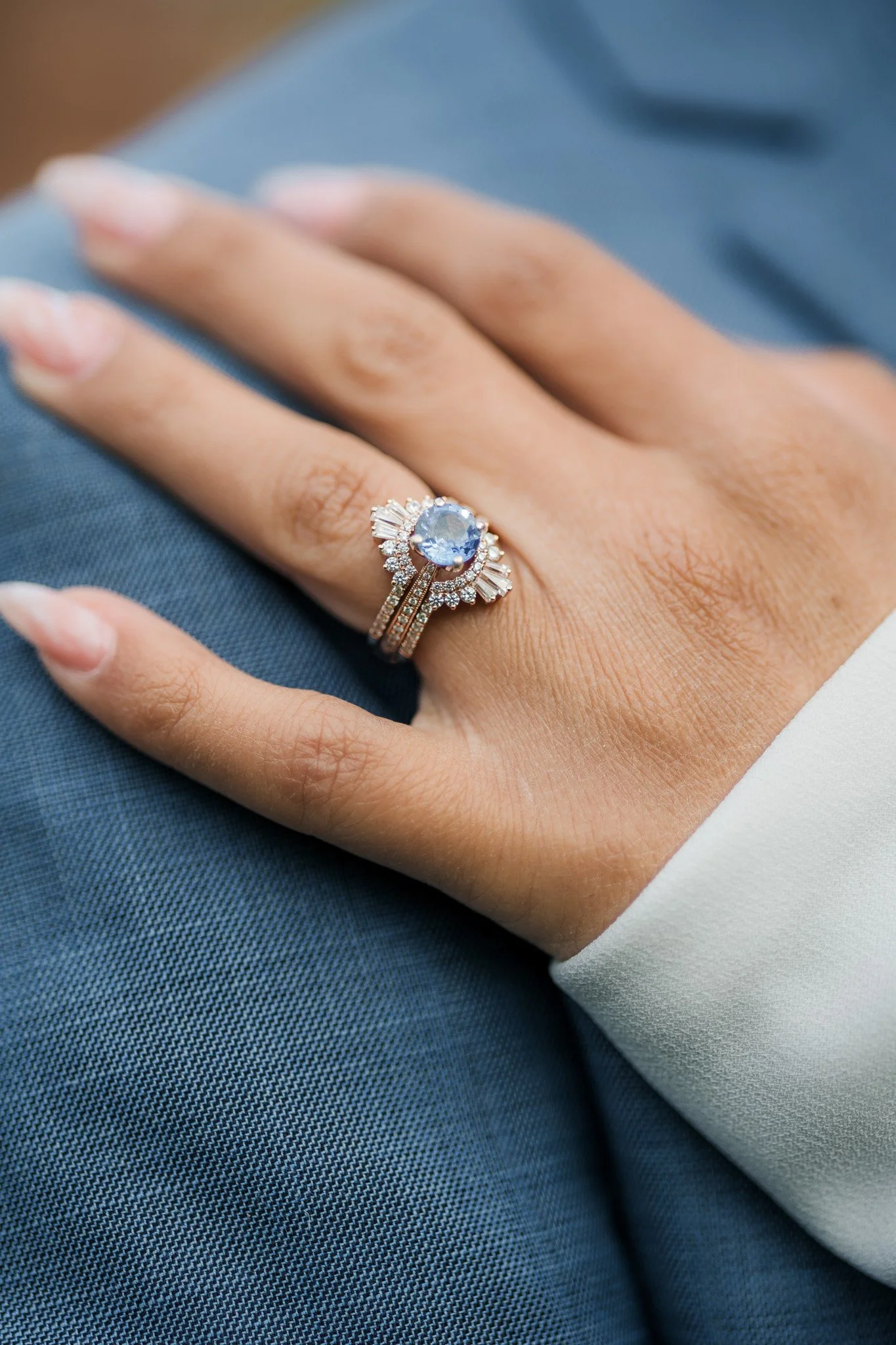 Close-up of a woman's hand showing an elegant engagement ring with a large central blue gemstone surrounded by small diamonds, and a wedding band with small diamonds, resting on a person's lap.