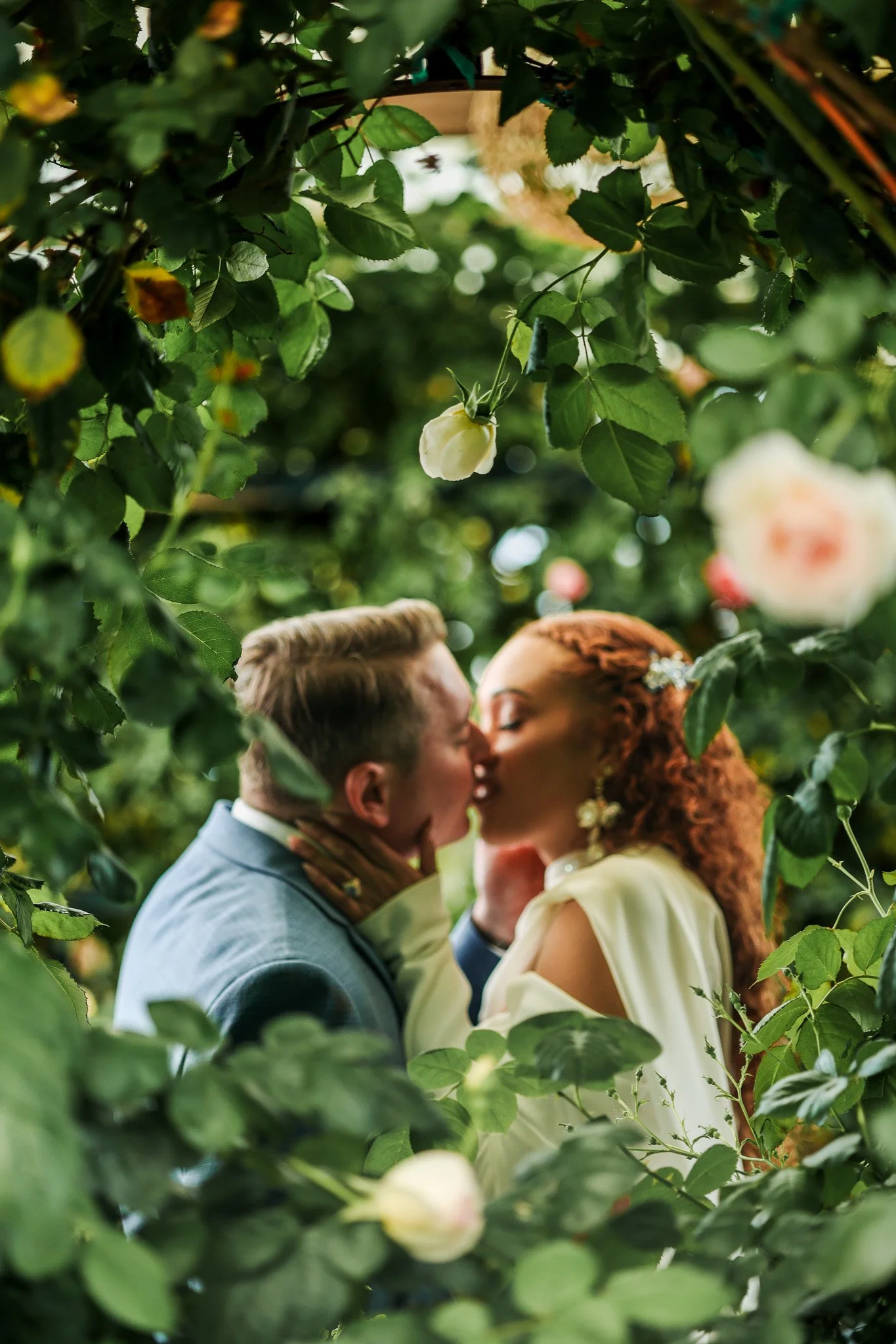 A couple sharing a kiss amid lush green foliage and blooming roses.