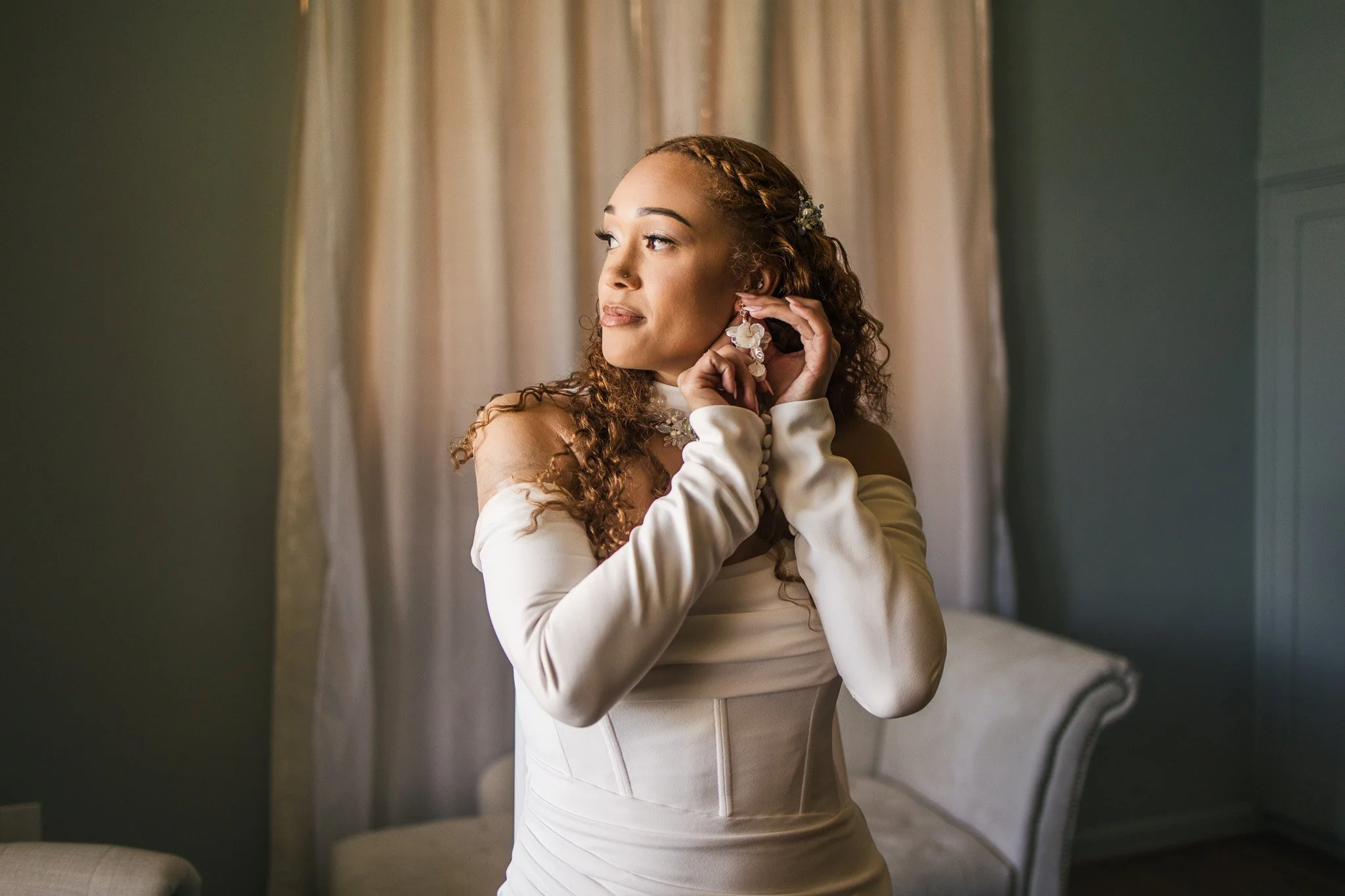 A bride adjusting her earring while standing in a room with cream-colored curtains and a beige sofa.