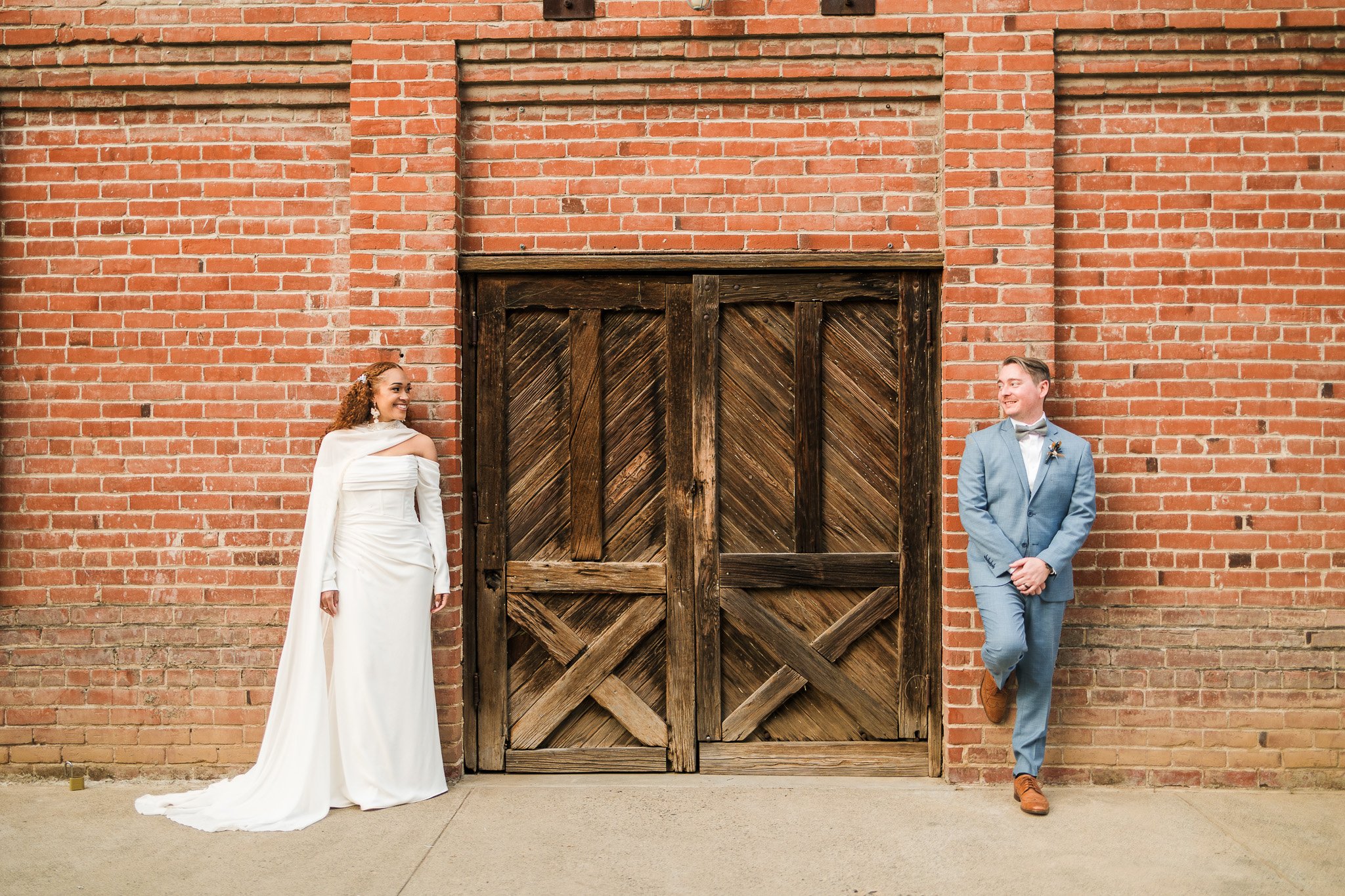 Bride and groom smiling at each other against a brick wall with a wooden door