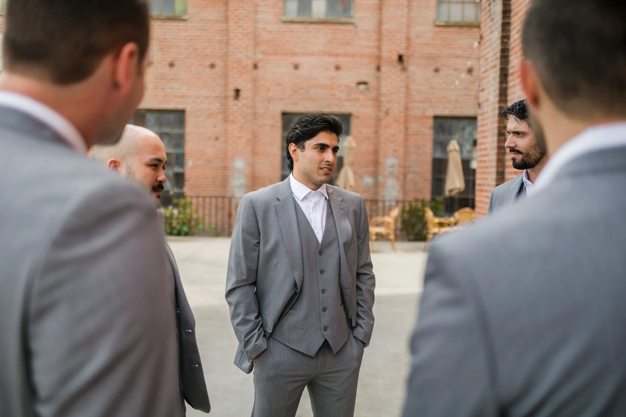A group of five men in suits having a discussion outside in an urban setting with a brick building in the background.