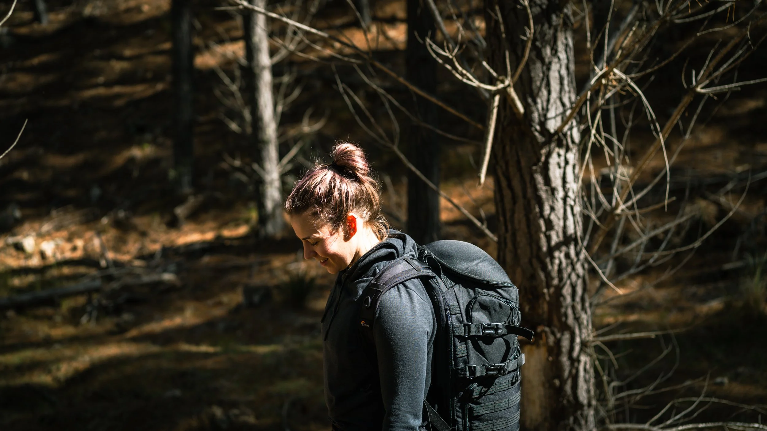 A woman with a bun hairstyle wearing a dark jacket and backpack, smiling as she walks through a wooded area with tall trees and sunlight filtering through the branches.