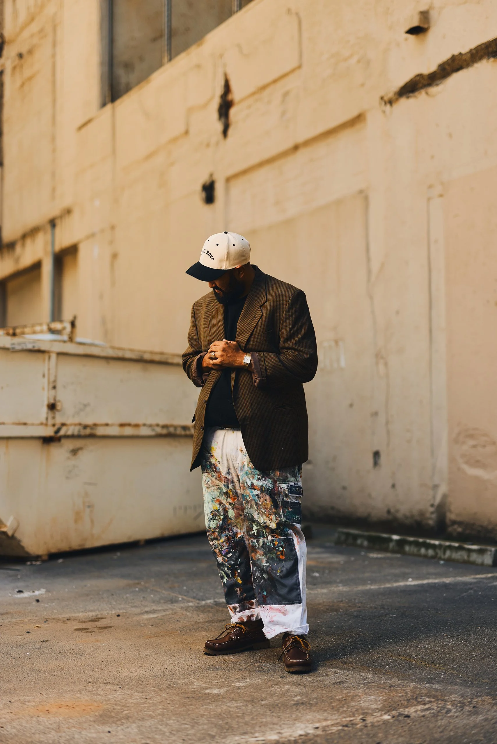 A man wearing a white baseball cap, sunglasses, a brown blazer, a black shirt, and paint-splattered pants stands in an alleyway with his head bowed and hands clasped.