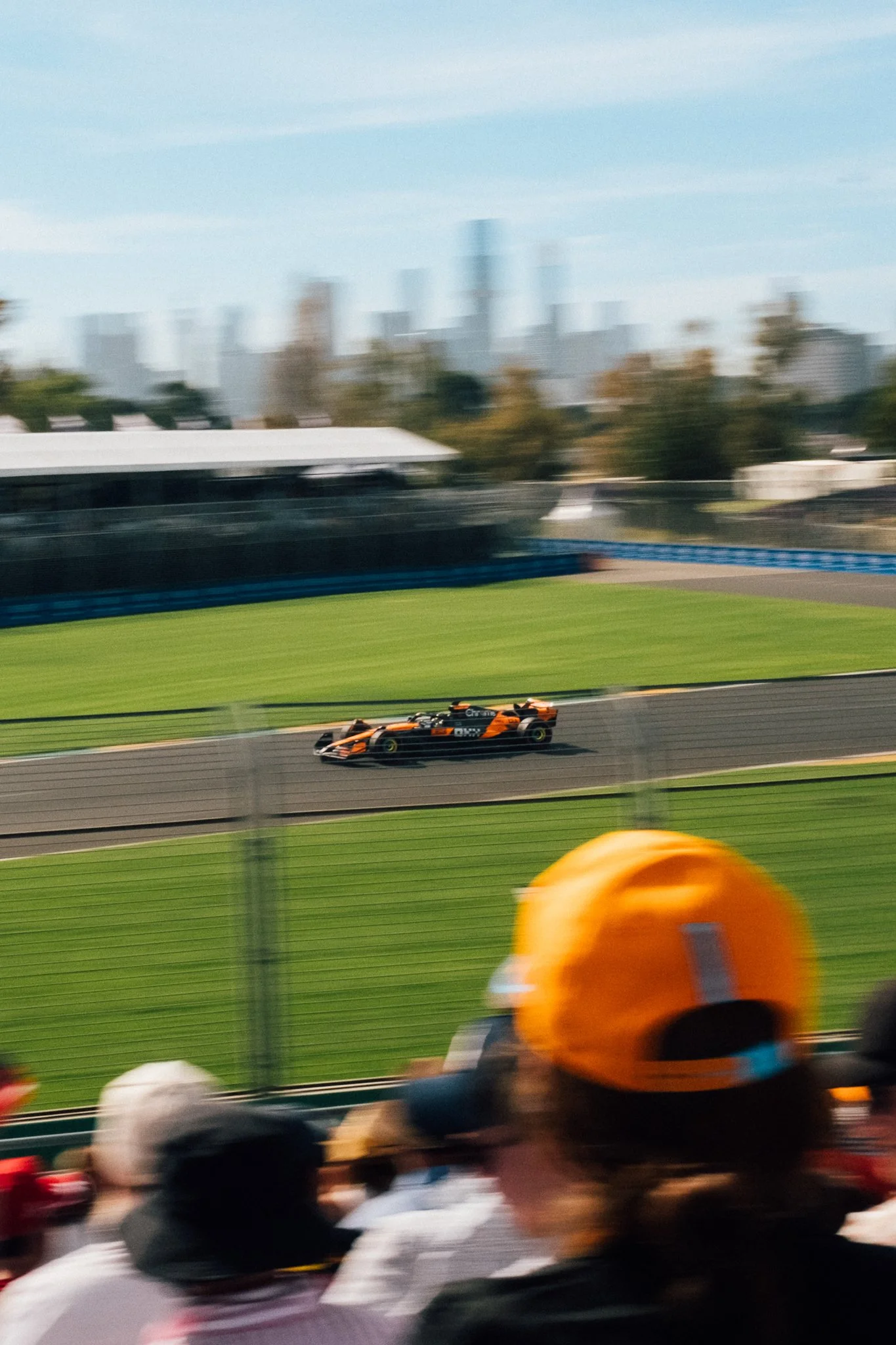 A race car on a track during a race, with spectators watching in the foreground, and a city skyline faintly visible in the background.