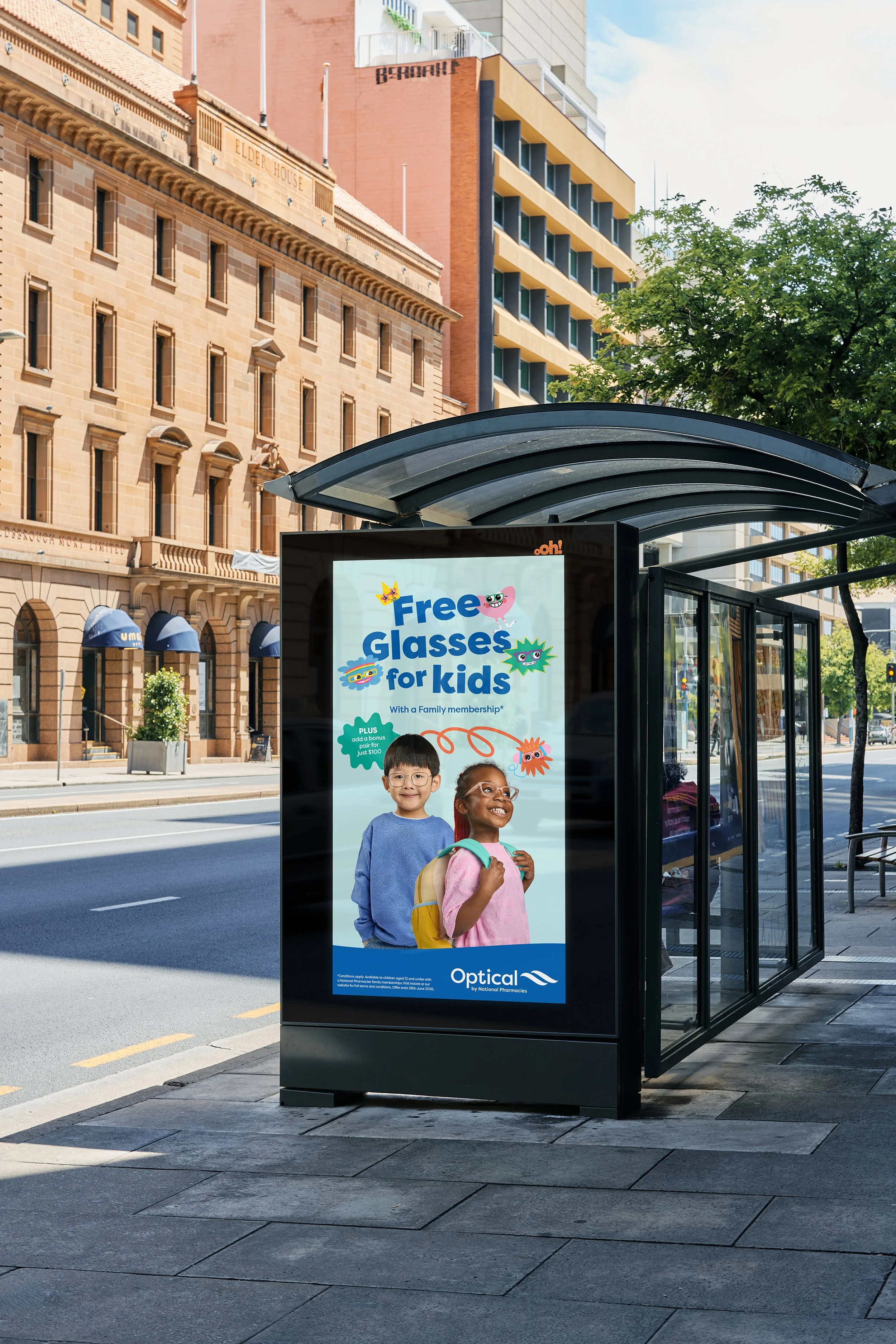 Advertisement at a bus stop for free glasses for kids, showing two children with backpacks and smiling. Urban street scene with buildings and trees in the background.