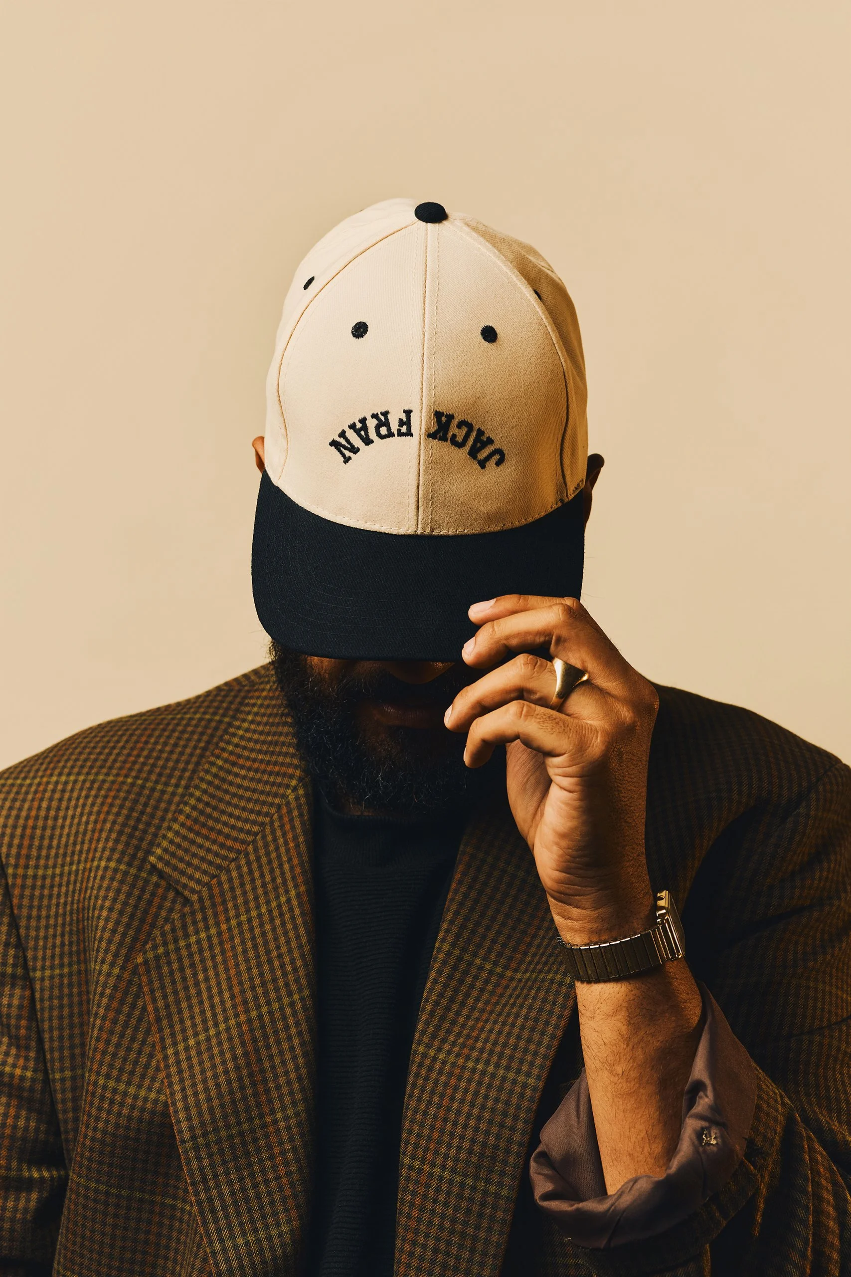 Man wearing a brown checkered blazer and black shirt, with a beige and black cap that says 'DARK LEAN', holding the brim with his right hand and partially covering his face, against a plain beige background.