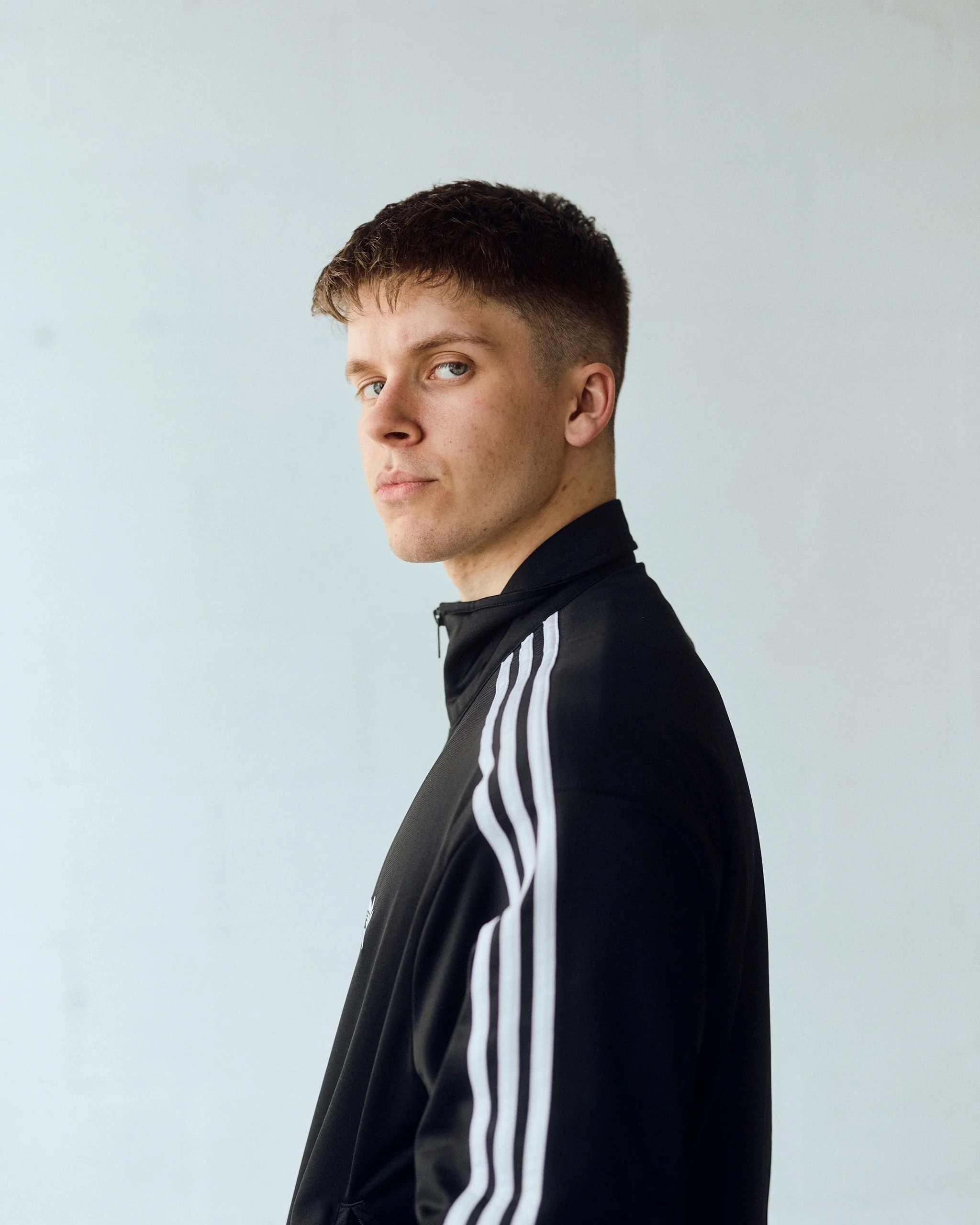 A young man with short brown hair and blue eyes wearing a black athletic jacket with white stripes on the sleeves, looking over his shoulder at the camera against a plain light gray background.