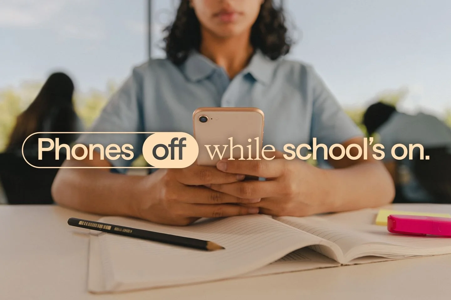 A student sitting at a desk with open notebook, pen, and highlighters, holding a smartphone. Text overlay reads 'Phones off while school's on'.