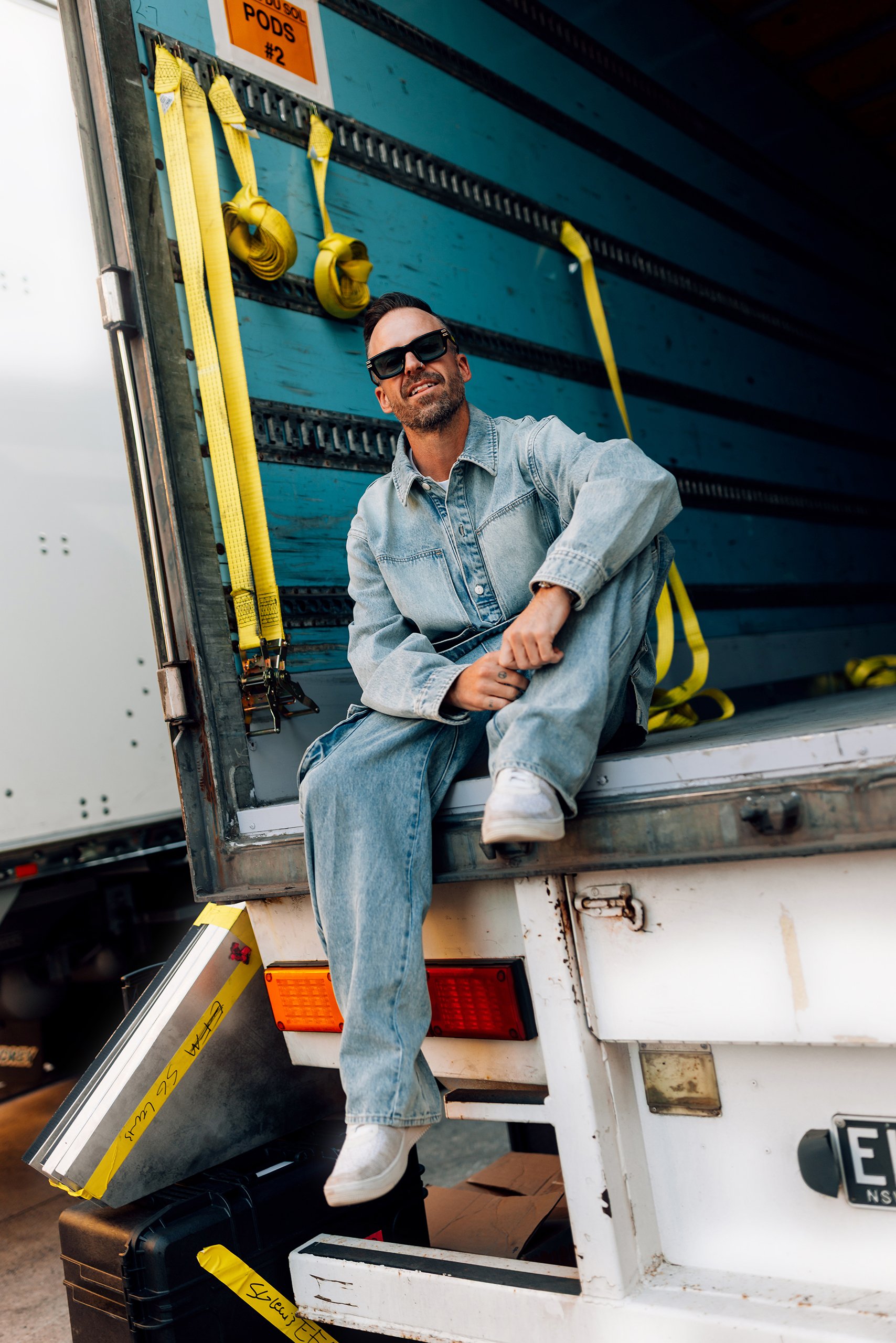 A man sitting on the back of a delivery truck with yellow straps hanging behind him, wearing sunglasses, denim jacket, and jeans, truck open and showing the cargo area.