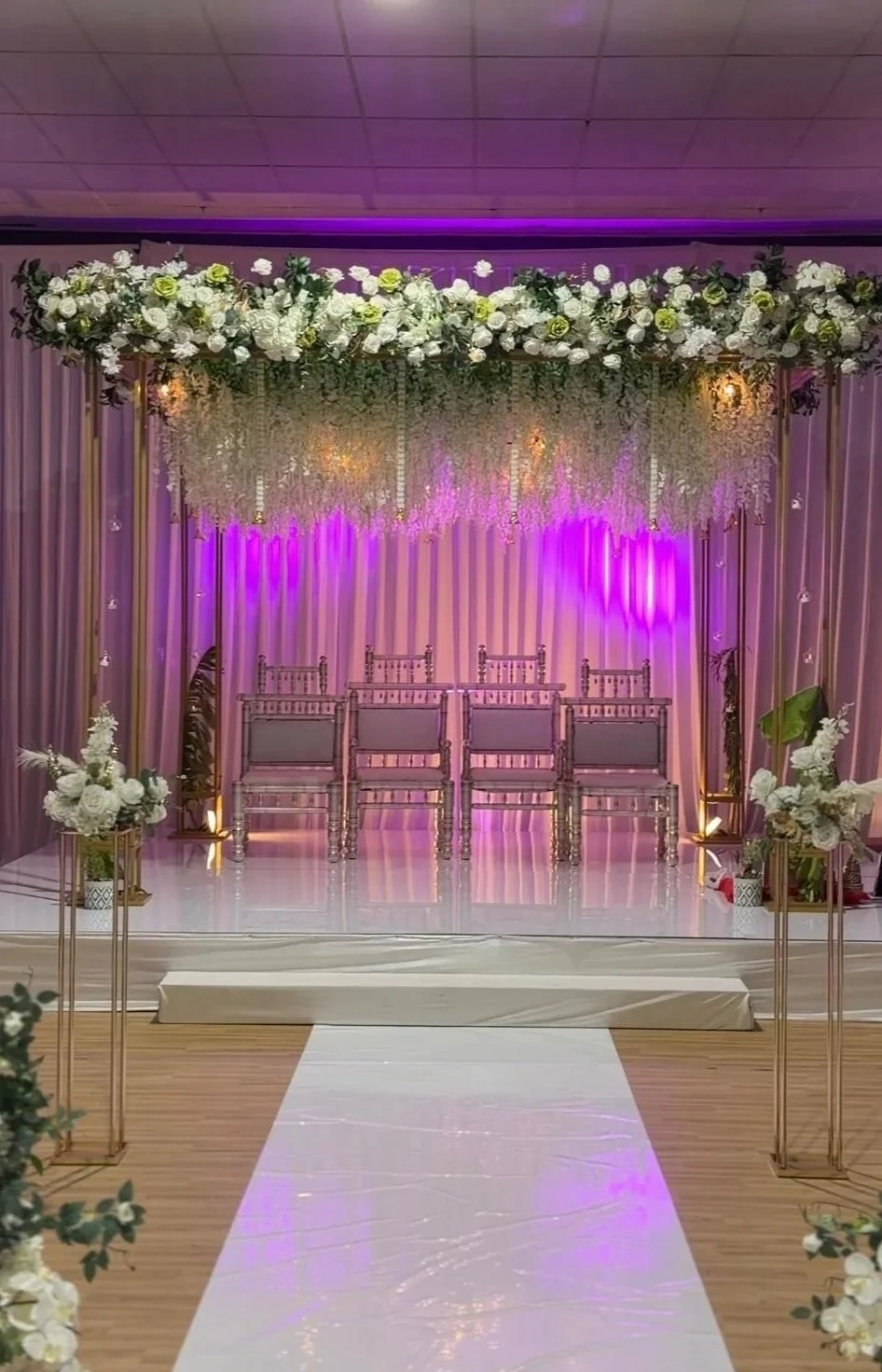 Wedding ceremony stage decorated with white flowers, purple lighting, and elegant chairs on a white glossy floor with floral arrangements on golden stands.