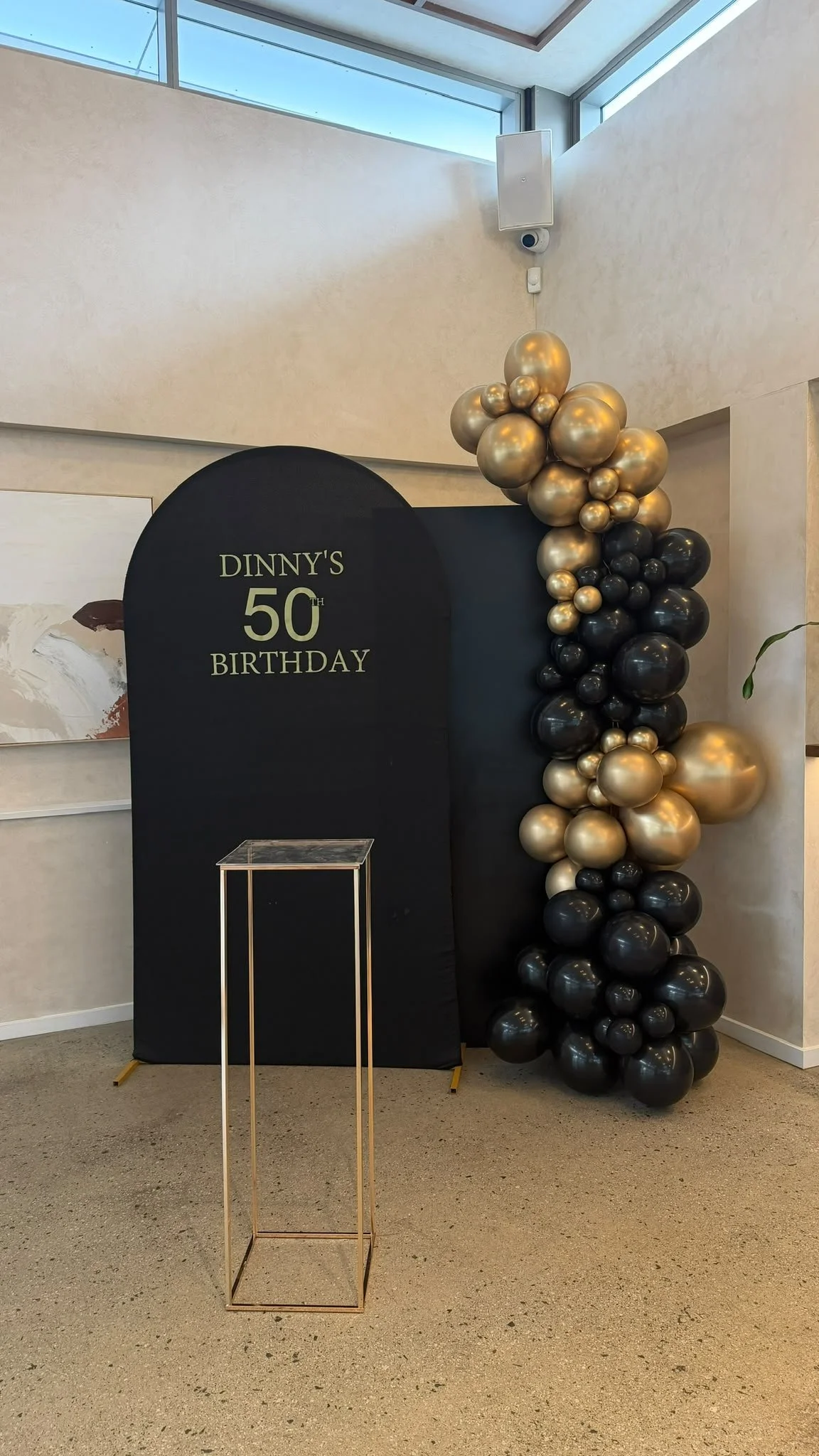A black backdrop with gold and black balloons forming a vertical arrangement. A sign on the backdrop reads "Dinnys 50th Birthday." An empty gold metallic stand is in front of the backdrop. The setting appears to be indoors with neutral-colored walls and a carpeted floor.