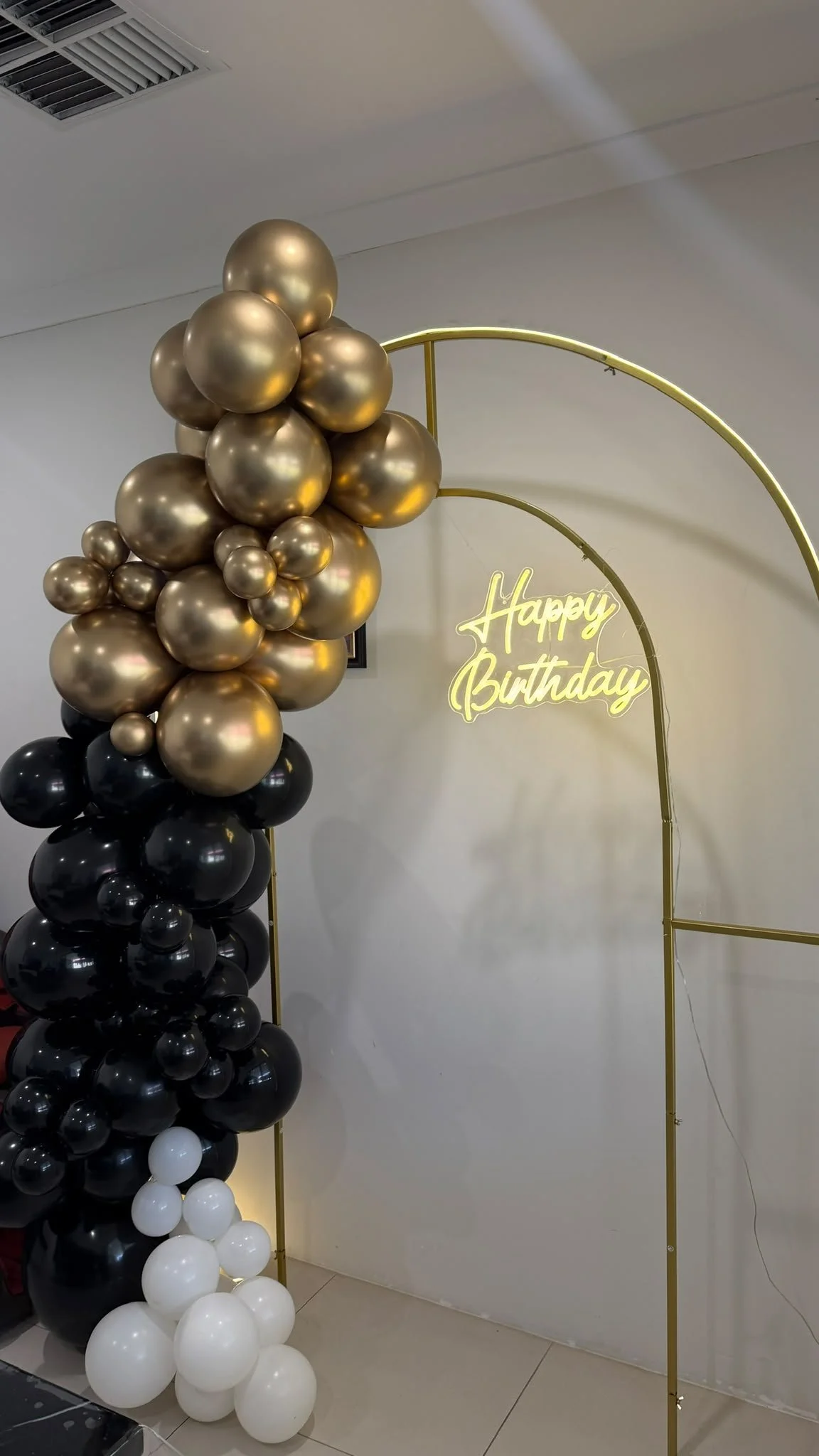 A balloon arrangement with black, white, gold, and bronze balloons next to a gold arch and a neon sign that says 'Happy Birthday' in yellow.