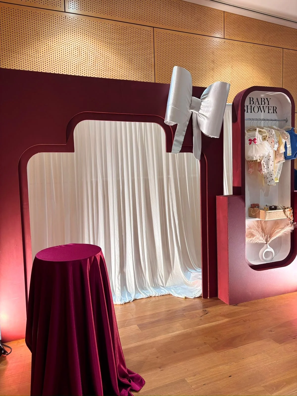 A photo of a gift wrapping station with a red and white theme. It features a white curtain backdrop, a round table with a red cloth, and a display labeled 'Baby Shower' showcasing baby clothes and accessories, with decorative elements like a large white bow and a pink pampas grass arrangement.