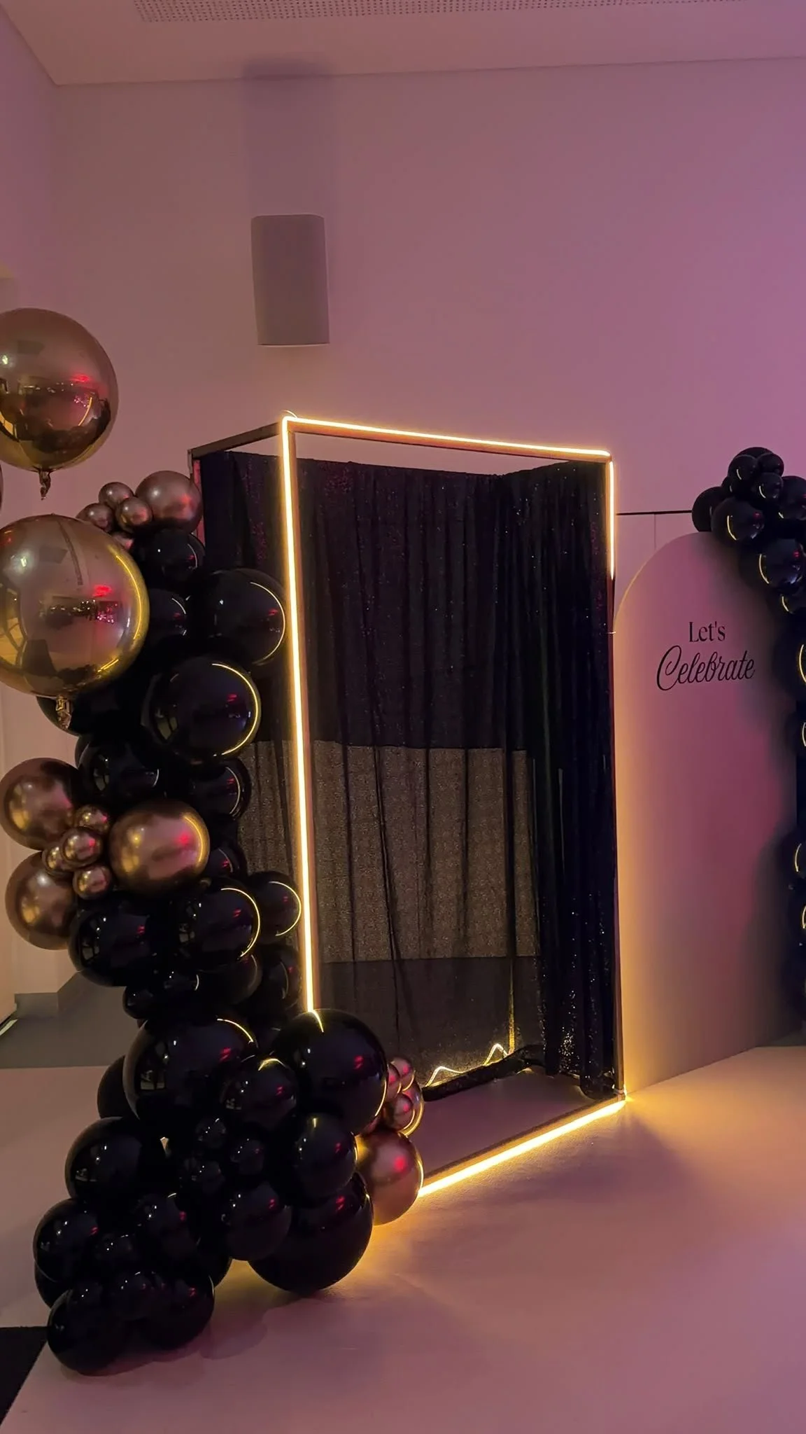 A photo booth setup with black and gold balloons on each side, a black curtain backdrop, and a rectangular light frame with a neon glow. There is a sign that says 'Let's Celebrate' on the right side.