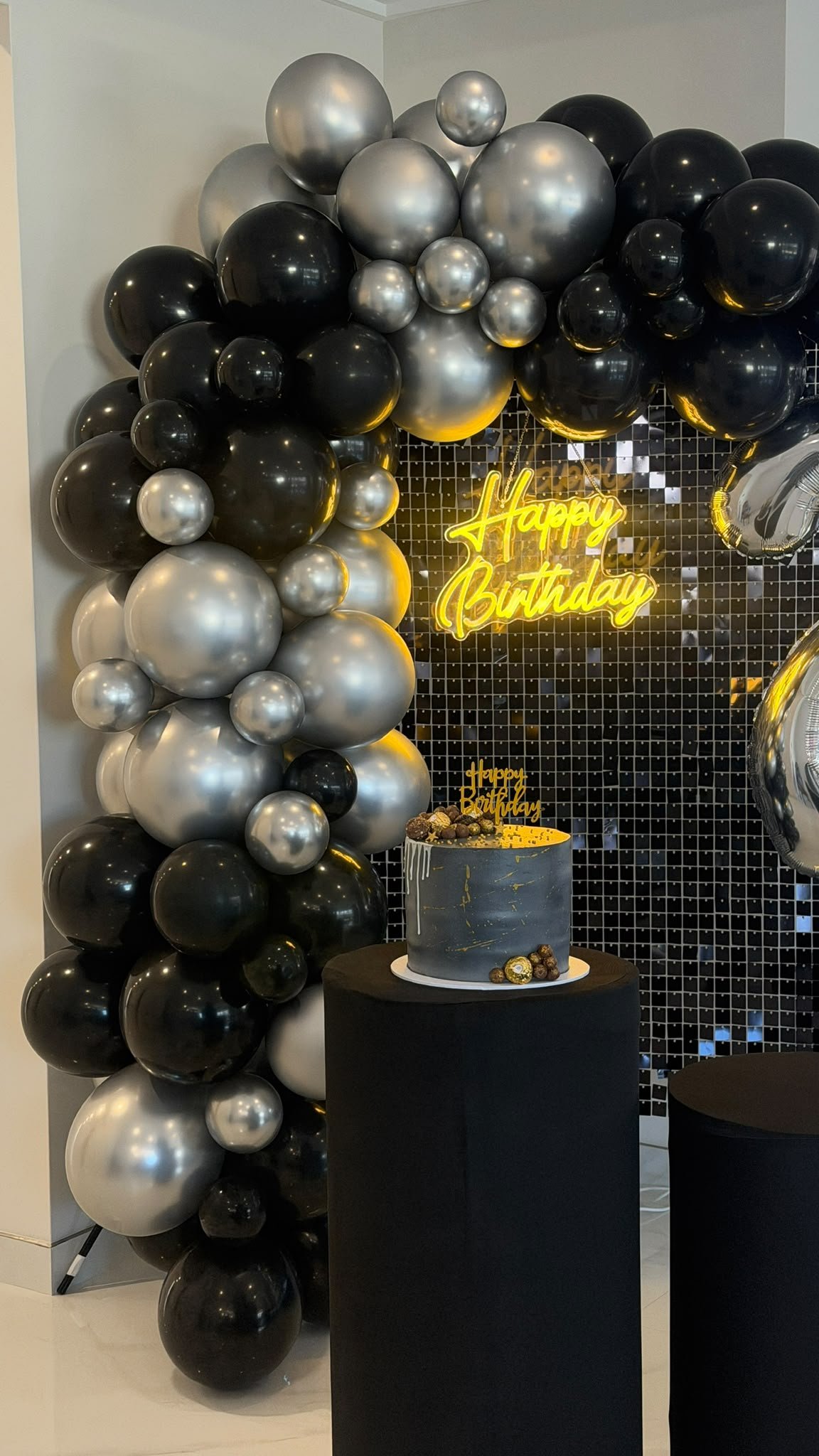 A birthday celebration setup with a black and gray theme. There is a cluster of black, gray, and silver balloons on the left, a black table with a gray birthday cake decorated with gold chocolates and a topper that says 'Happy Birthday'. In the background, a neon yellow sign says 'Happy Birthday' on a black mosaic wall.