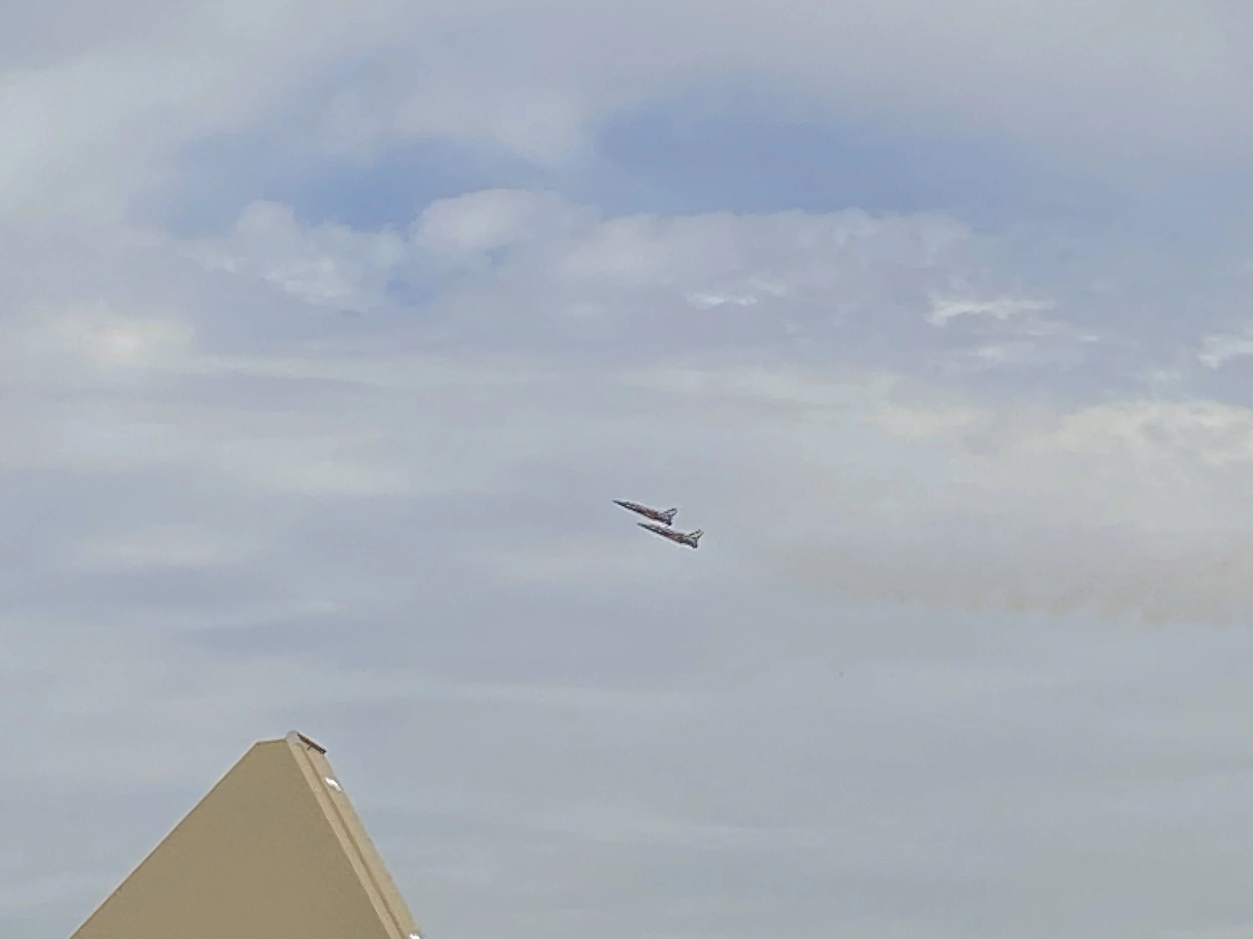 Two airplanes flying in the sky above a building with a sloped roof, with clouds in the background.