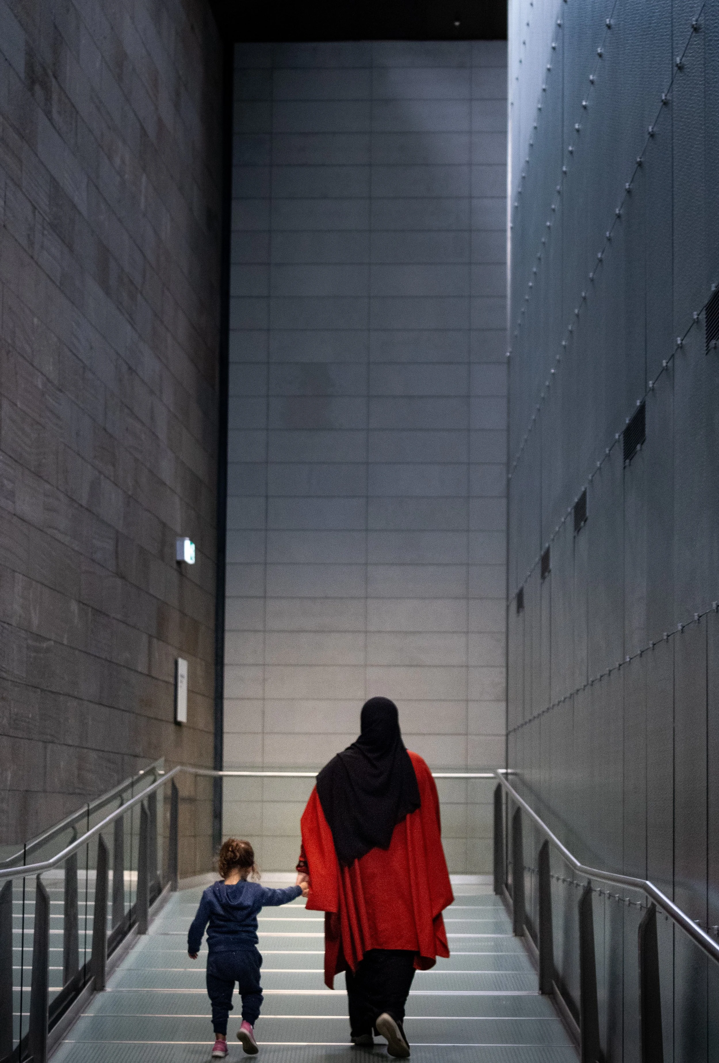 A woman wearing a black headscarf and a red coat holding the hand of a young girl walking up a staircase inside a modern building.
