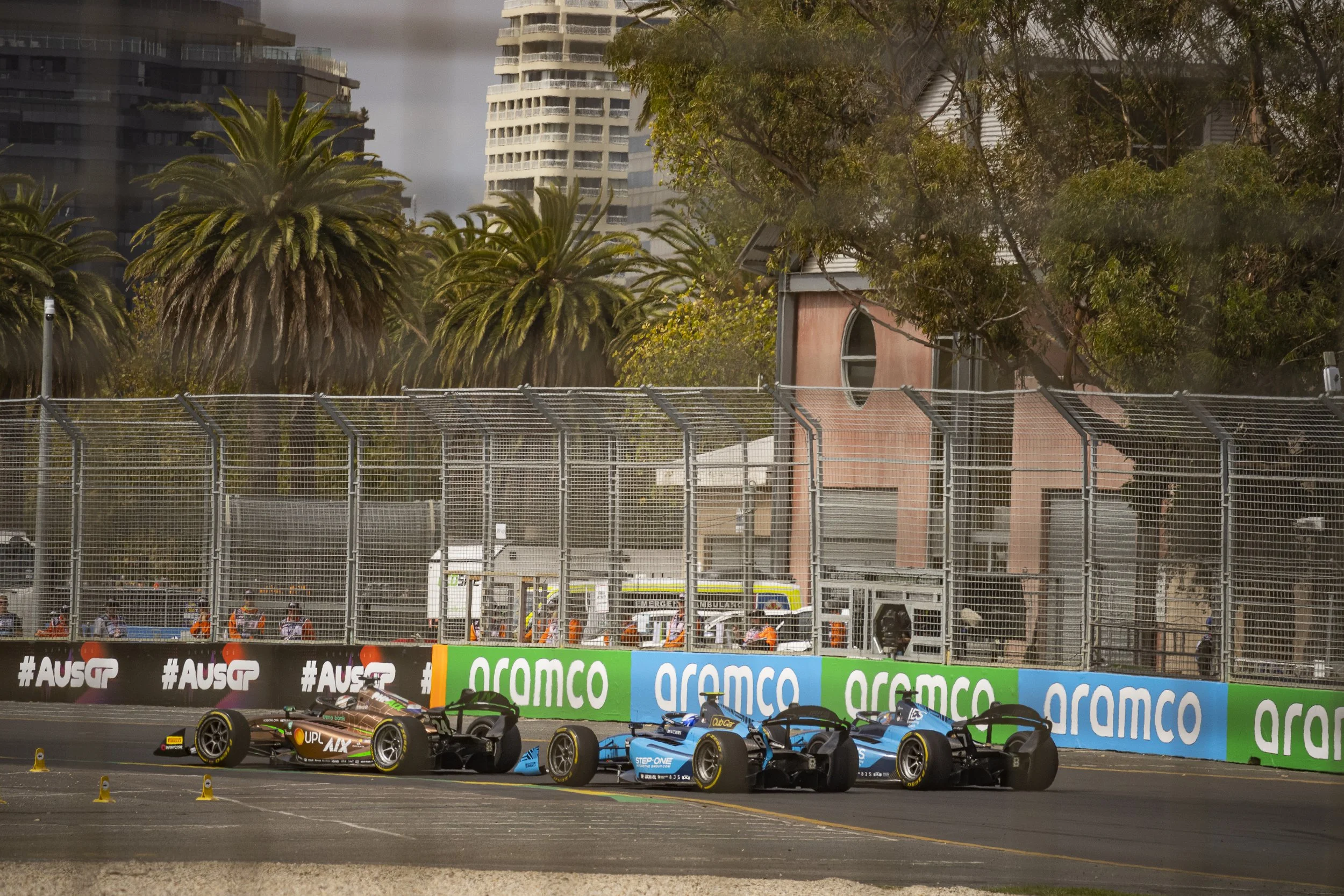 Three race cars on a track during a race, with a barrier, trees, and tall buildings in the background.