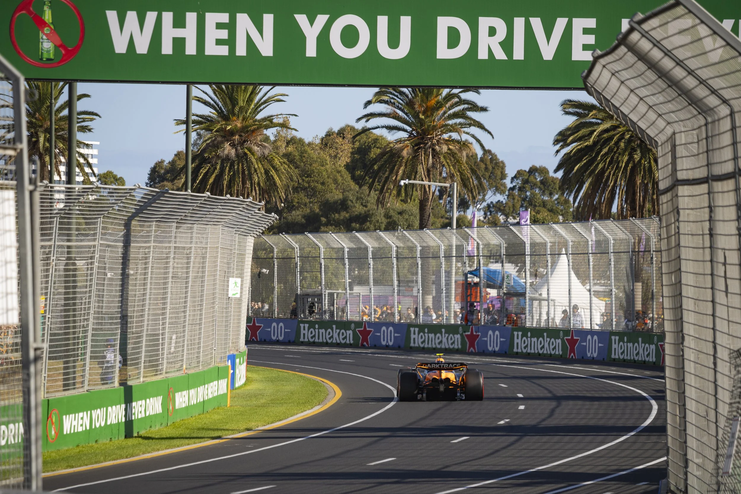 Formula 1 race car driving along a street circuit with safety barriers, palm trees, and advertising banners, under a large overhead sign that reads 'When You Drive'.