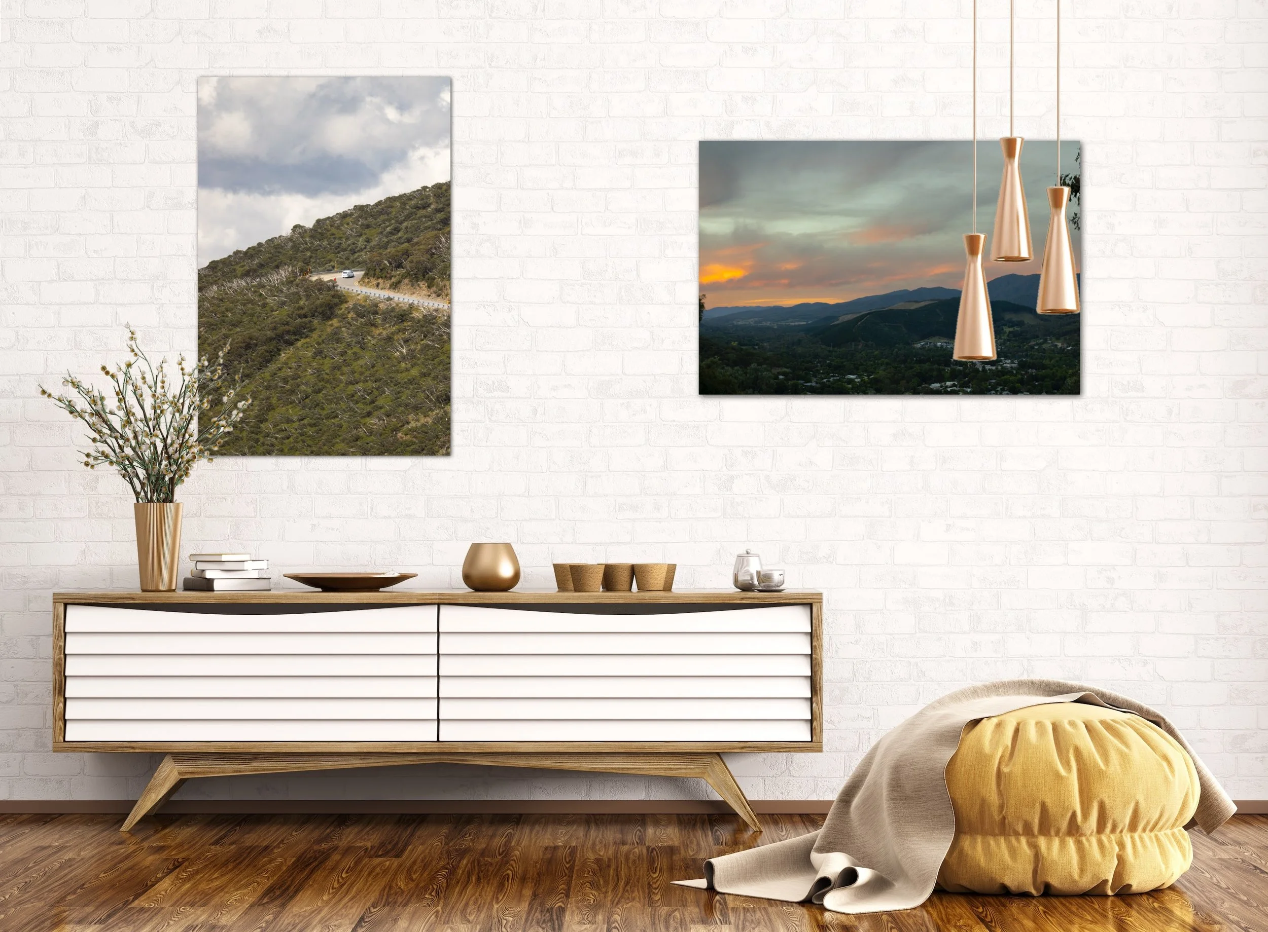 Modern interior with a wooden sideboard against a white brick wall, decorated with vases and books. Two framed landscape photographs hang on the wall: one with a mountain road and the other with a sunset over mountains. A yellow pouf and a blanket are on the wooden floor.
