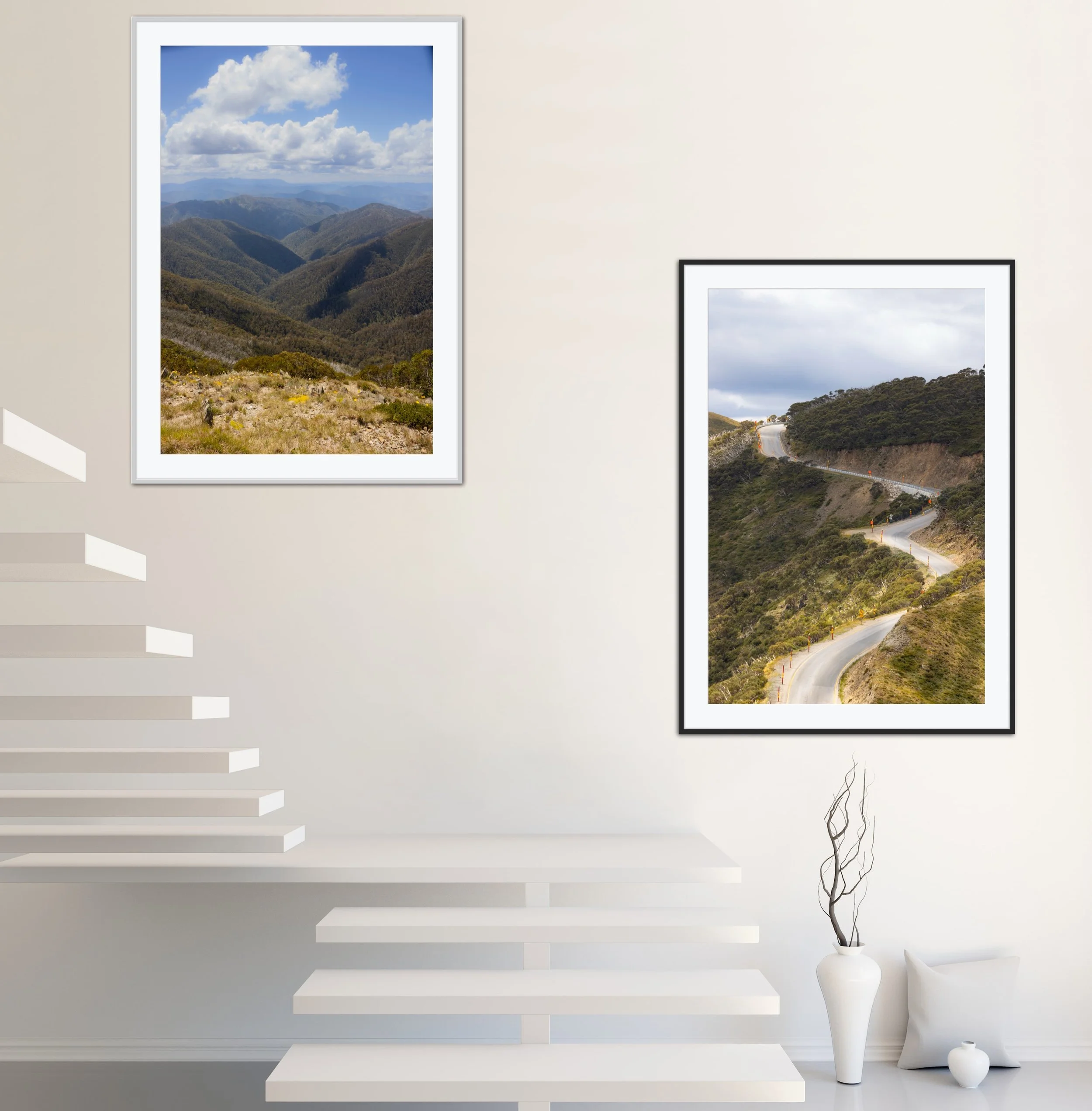 Interior of a modern home with white walls, featuring two framed landscape photographs of mountain scenery, a white vase with decorative twigs, and a white cushion on the floor.
